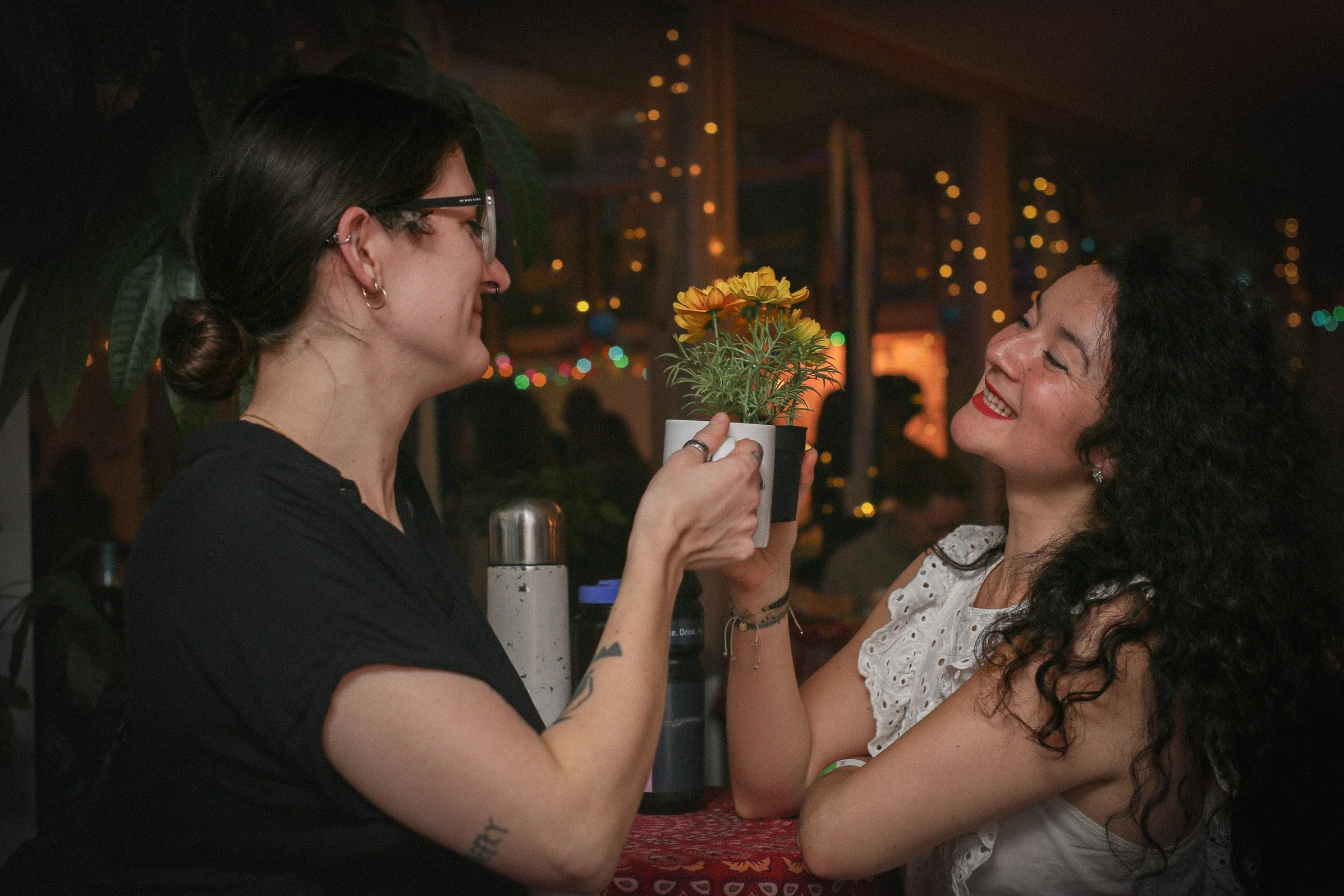 Two women sharing a moment at a bar or restaurant; one woman hands the other a potted plant with yellow flowers; the woman receiving the plant is smiling with curly black hair and white eyelet top; the woman giving the plant has glasses, earrings, dark hair in a bun, and tattoos.