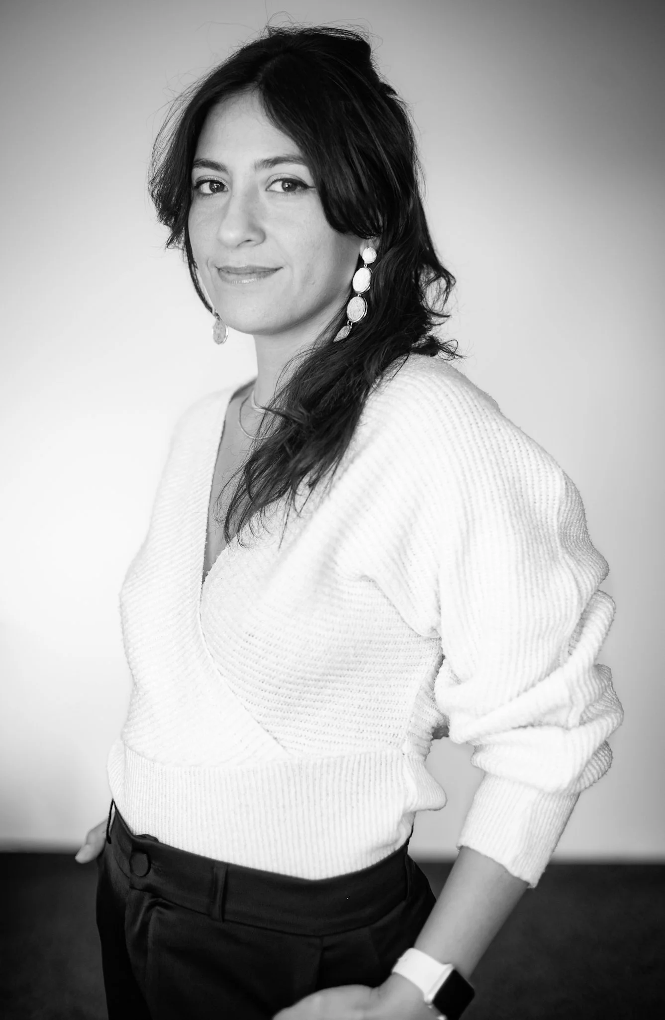 Black and white portrait of a woman with dark, wavy hair. She is wearing a light-colored, ribbed sweater and dark trousers, accessorized with long dangling earrings and a smartwatch. She stands with one hand in her pocket, looking confidently at the camera.