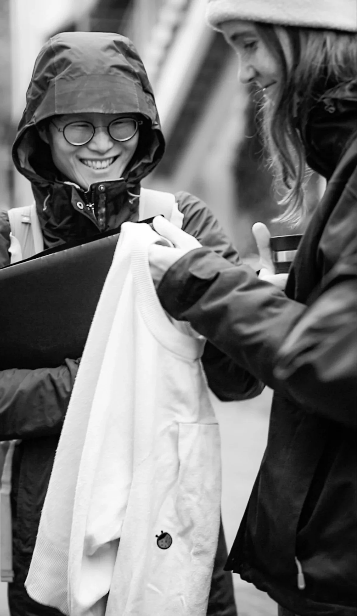Two women, one wearing glasses and a hooded jacket, smiling, exchanging a hoodie with a small ladybug pin, outdoors.