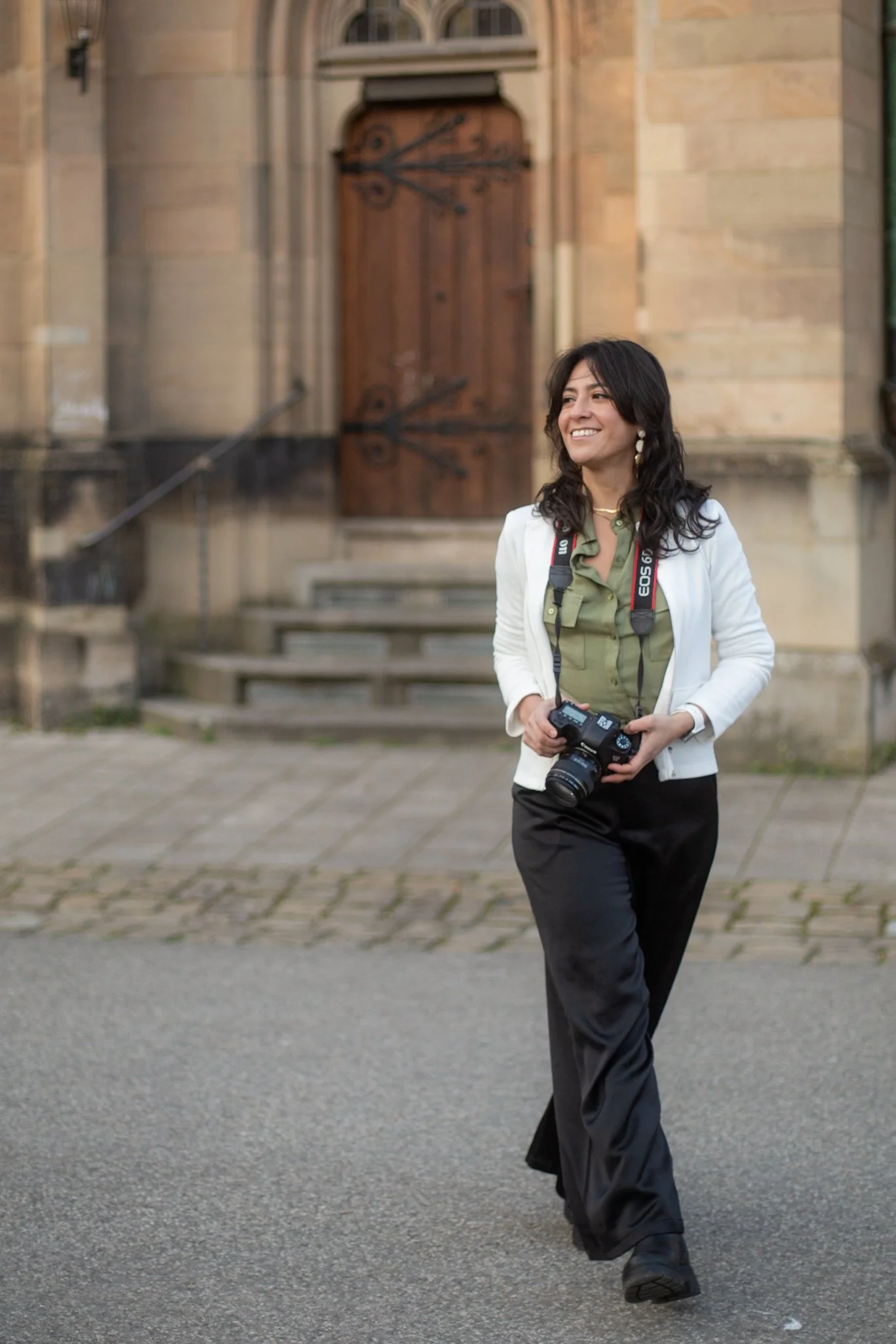 A woman with dark hair, smiling, holding a camera, walking outside in front of a stone building with a wooden door.