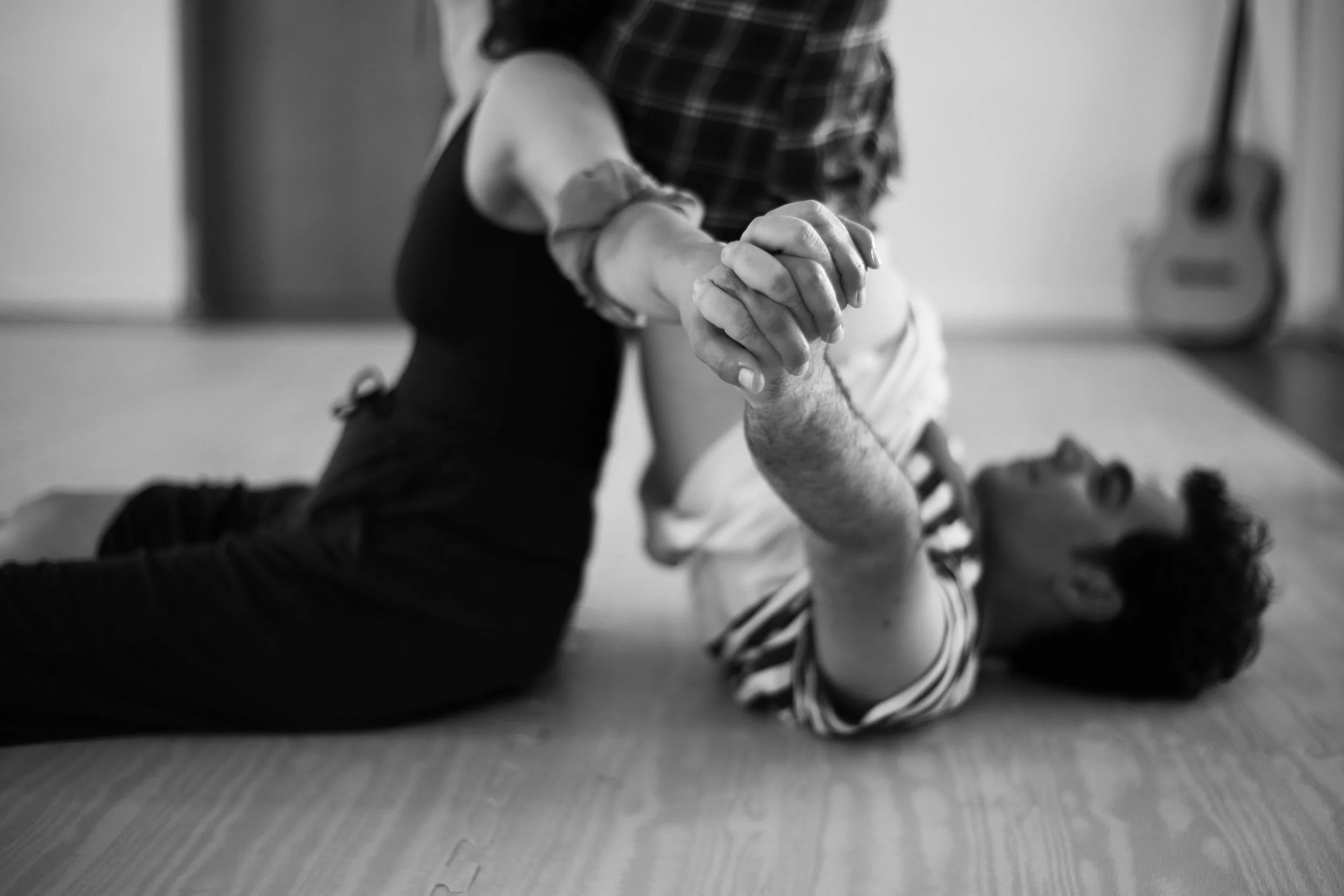 A person lying on their back on a wooden floor, holding hands with another person who is kneeling and leaning over them. A guitar is visible in the background.