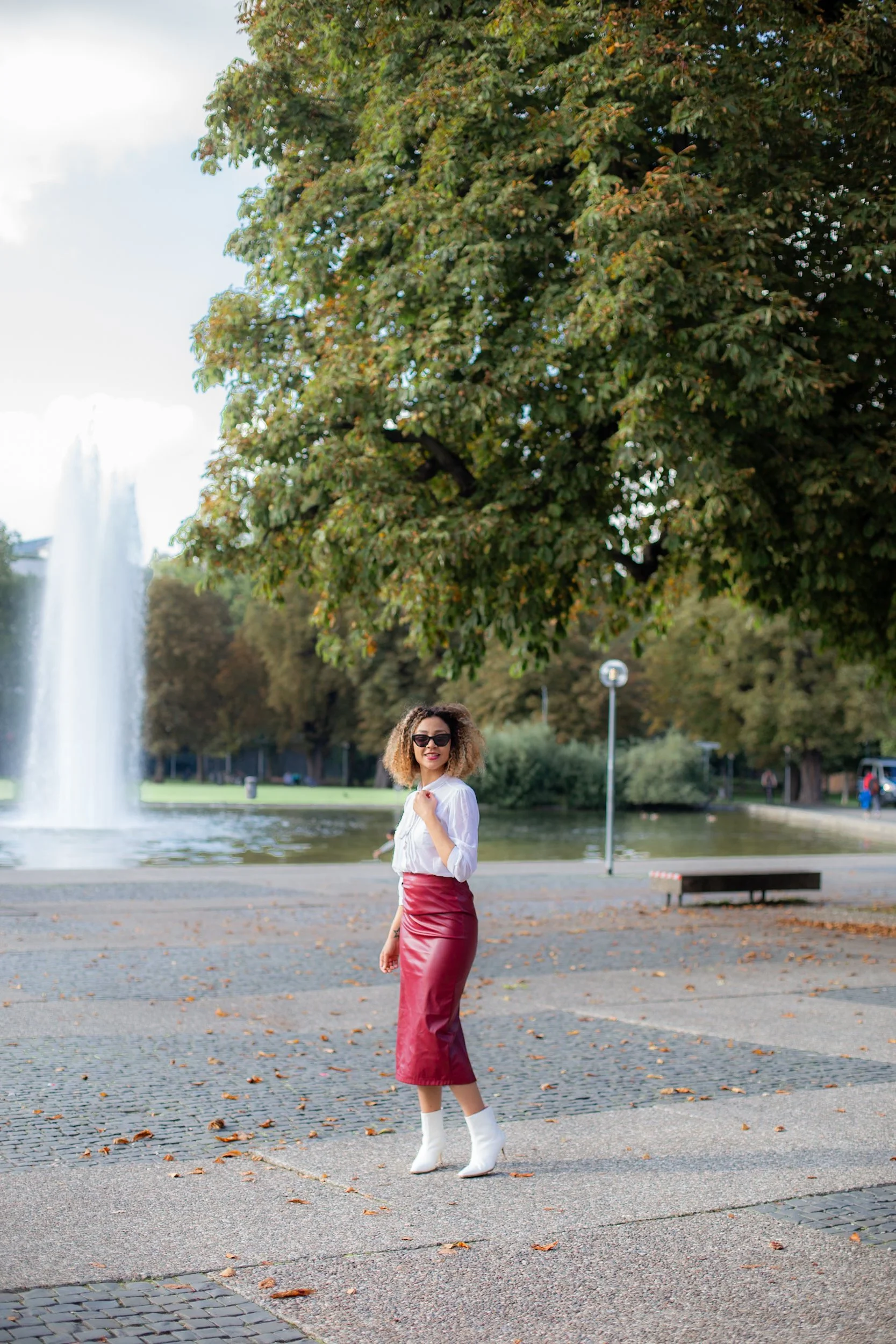 A woman stands in a park near a fountain, wearing a white blouse, red leather skirt, white boots, and sunglasses, smiling at the camera.