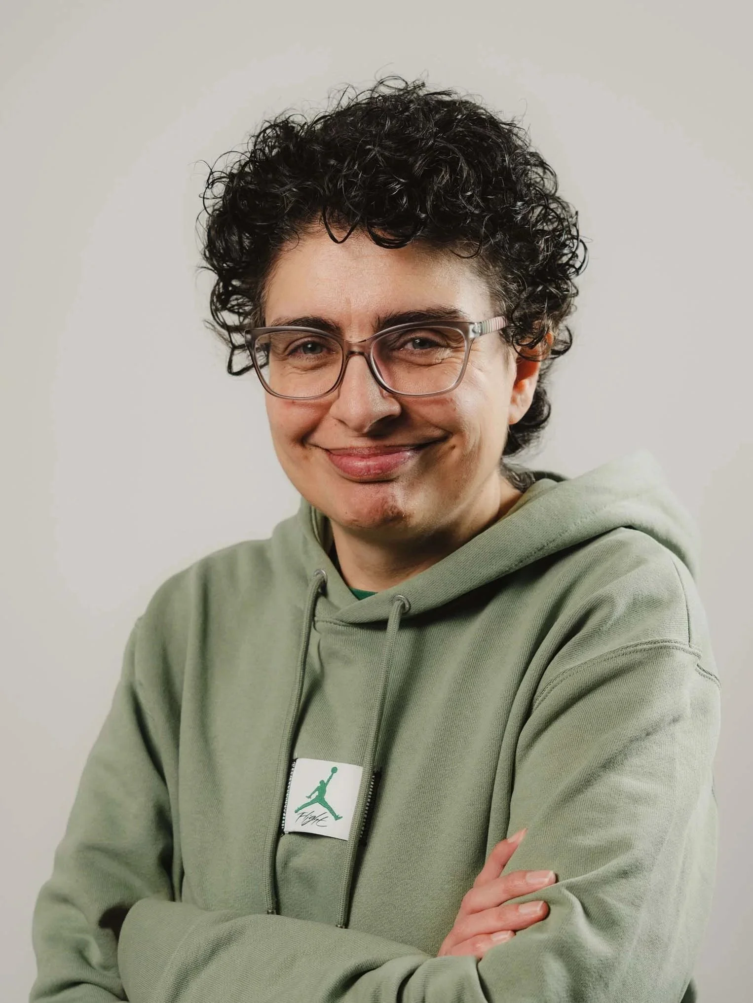 Close-up of a smiling woman with short curly hair wearing glasses and a green hoodie, standing against a plain neutral background.
