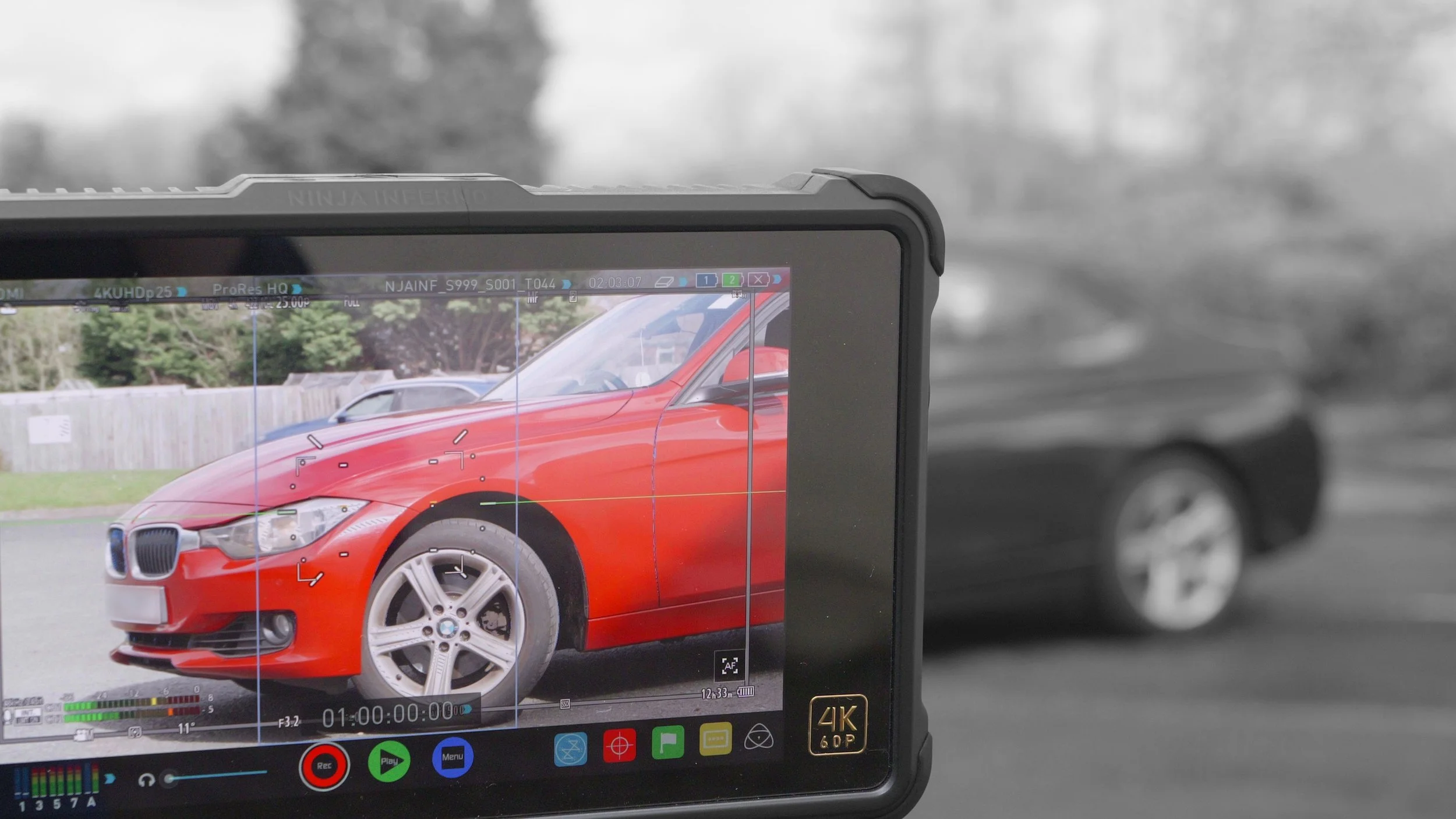 The image shows a camera or recording device capturing a red BMW car with a black grille and silver wheels, with a blurred background of trees and a fence.