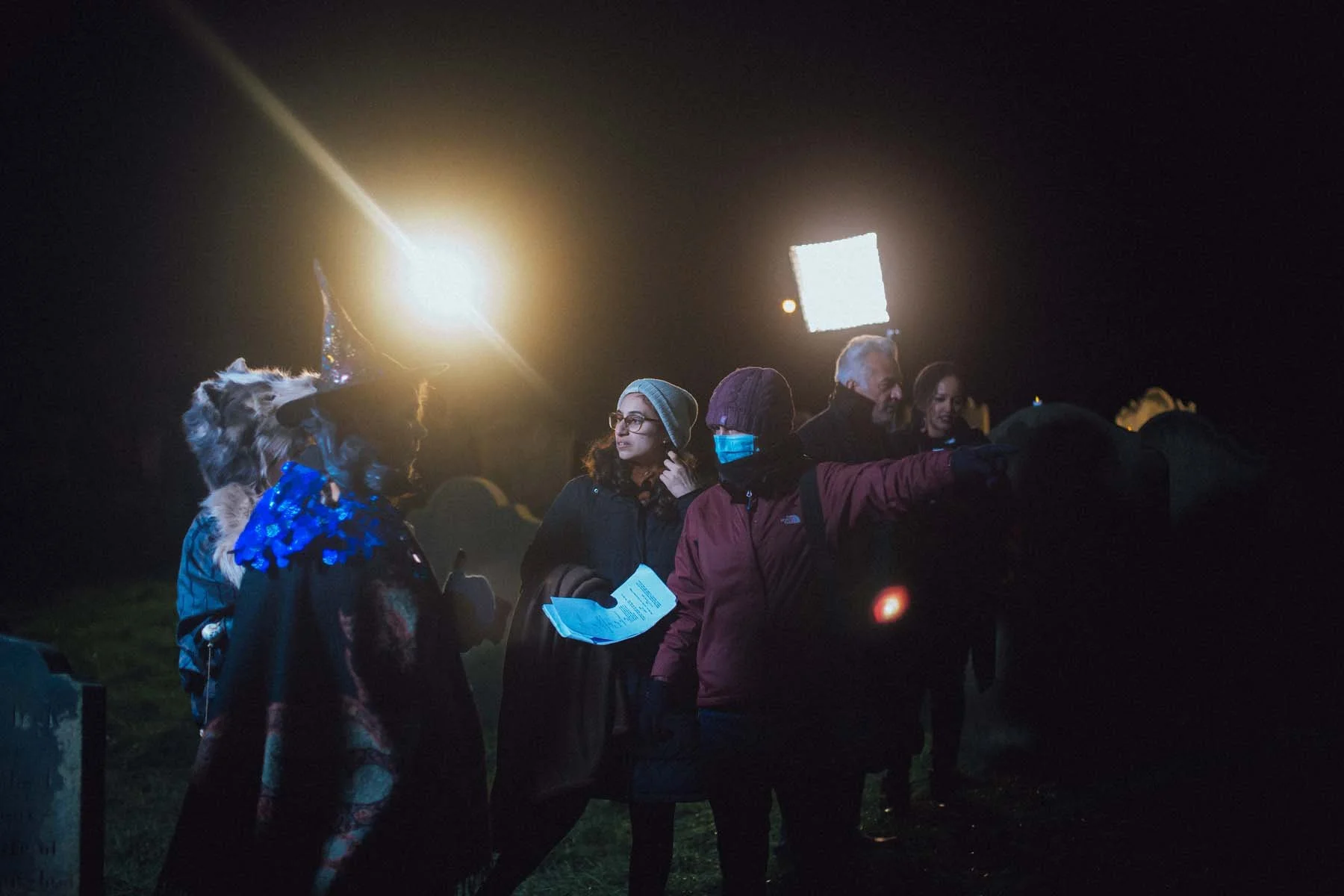 People gathered outdoors at night, some wearing masks and winter clothing, with bright lights and a dark sky in the background.