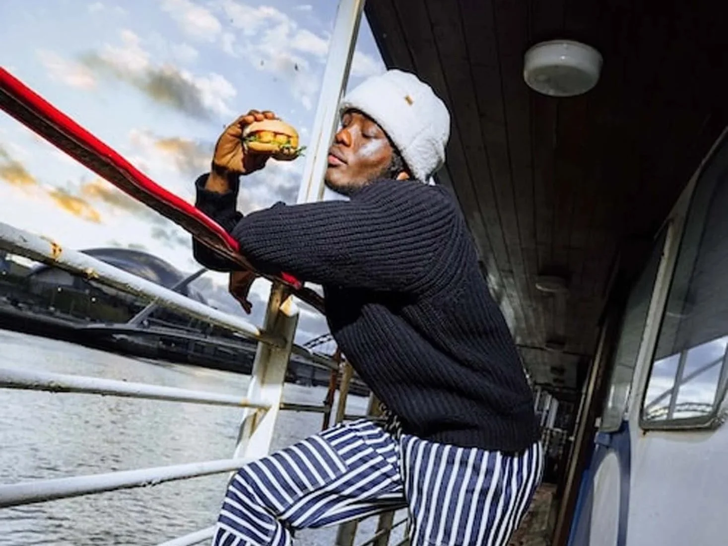 A man with dark skin, wearing a gray beanie, black sweater, and striped pants, leans on a boat's railing while eating a burger outdoors during daytime with a cloudy sky.