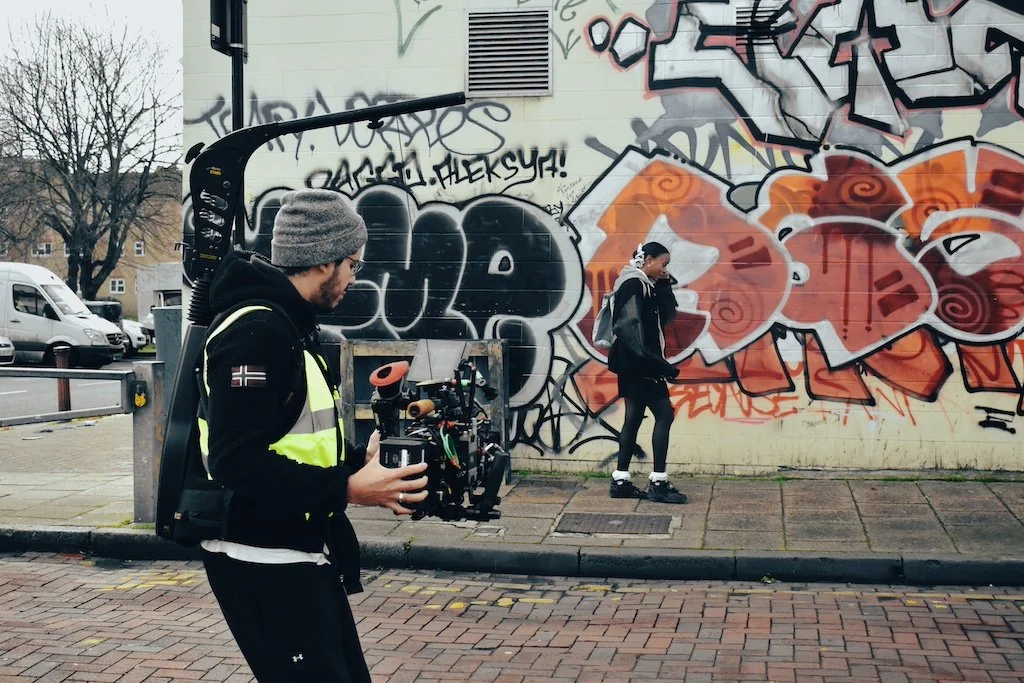 A man with a video camera and headphones filming a woman walking past graffiti on a wall.