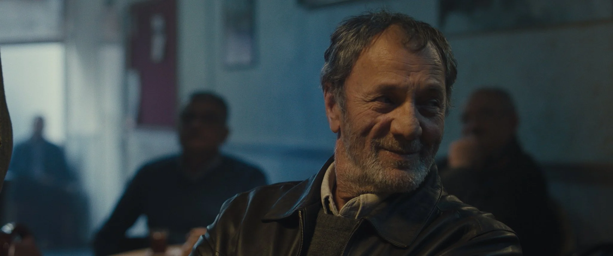 An older man with gray hair and a beard smiling in a dimly lit indoor setting, with blurred figures in the background.