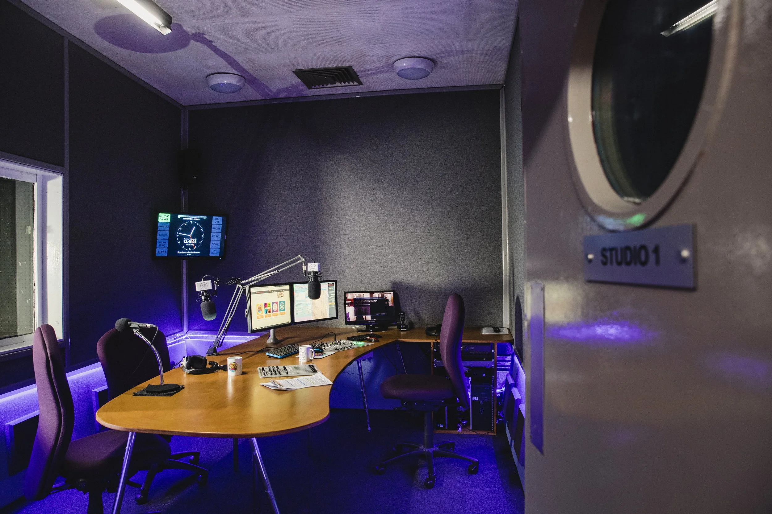 A radio studio with a table, chairs, microphones, and multiple computer monitors. The room has purple lighting and a clock on the wall reading 12:45.