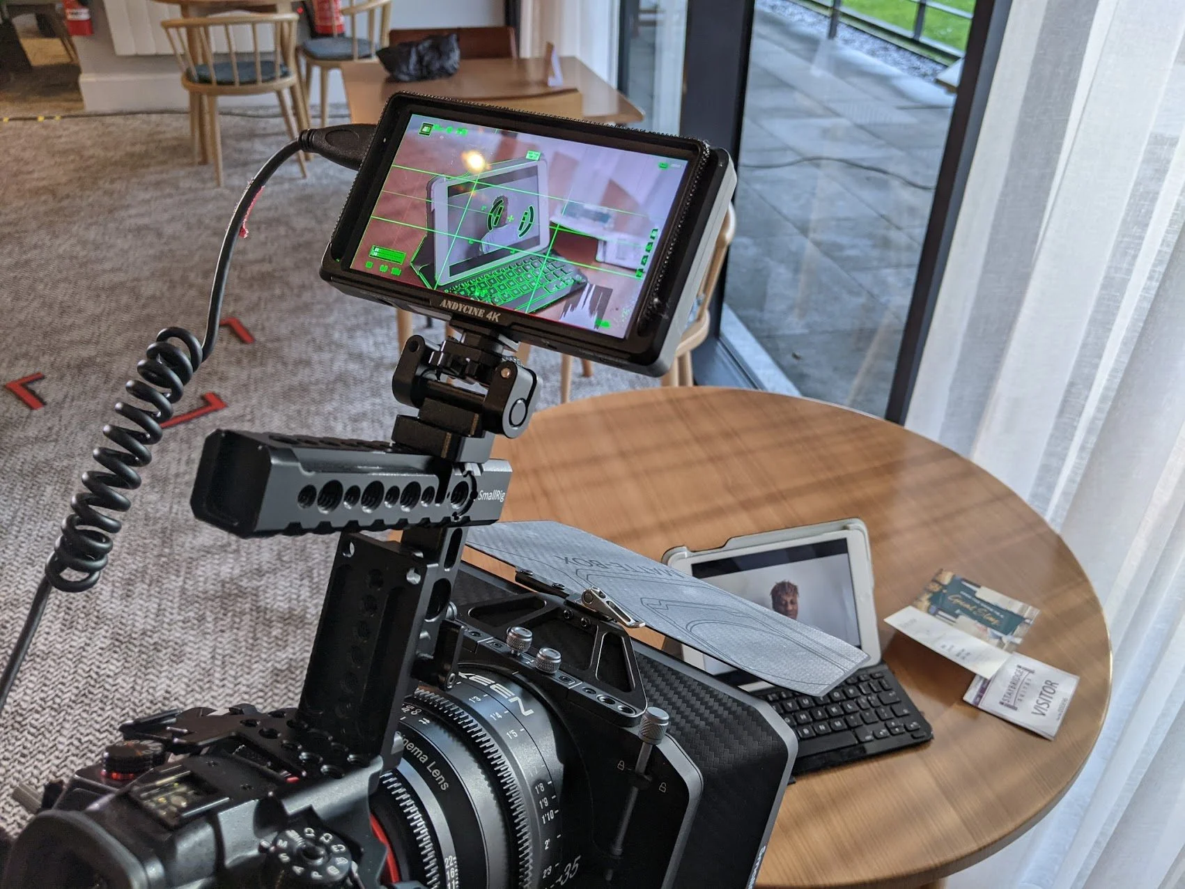 Camera mounted on a rig recording a video on a computer monitor, with a keyboard, papers, and a tablet on a wooden table in a room with a carpet and large windows.