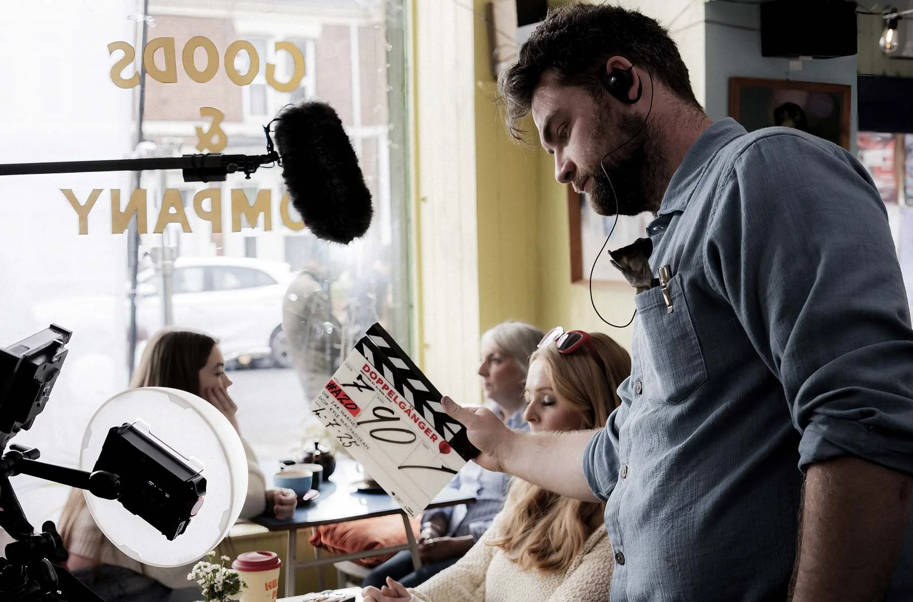 A man holding a clapperboard on a film set inside a cafe with people sitting at tables and a camera with a ring light in the foreground.