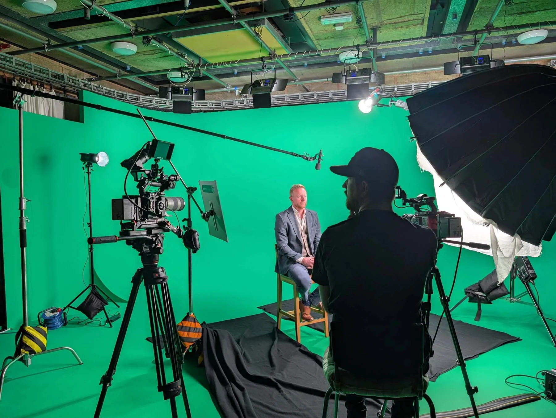 A video production studio with a green screen background. Two men are seated, one in a gray suit being recorded, and a crew member is operating camera equipment. Lighting and sound gear are set up around them.