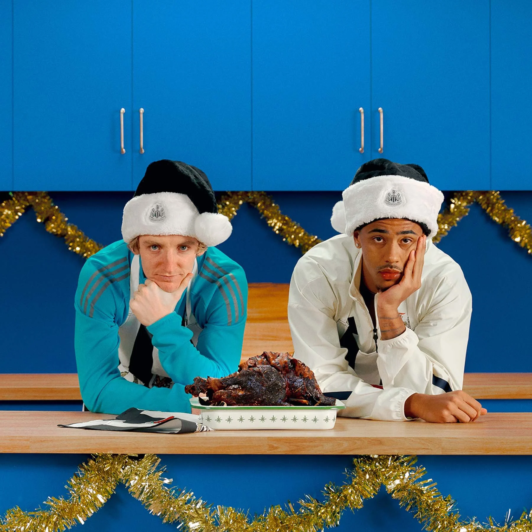 Anthony Gordon and Jacob Ramsey  wearing Santa hats sitting at a table in front of a blue wall with gold tinsel. They both have their chins resting on one hand and are looking at the camera. There is a roasted pig on a white platter in front of them.