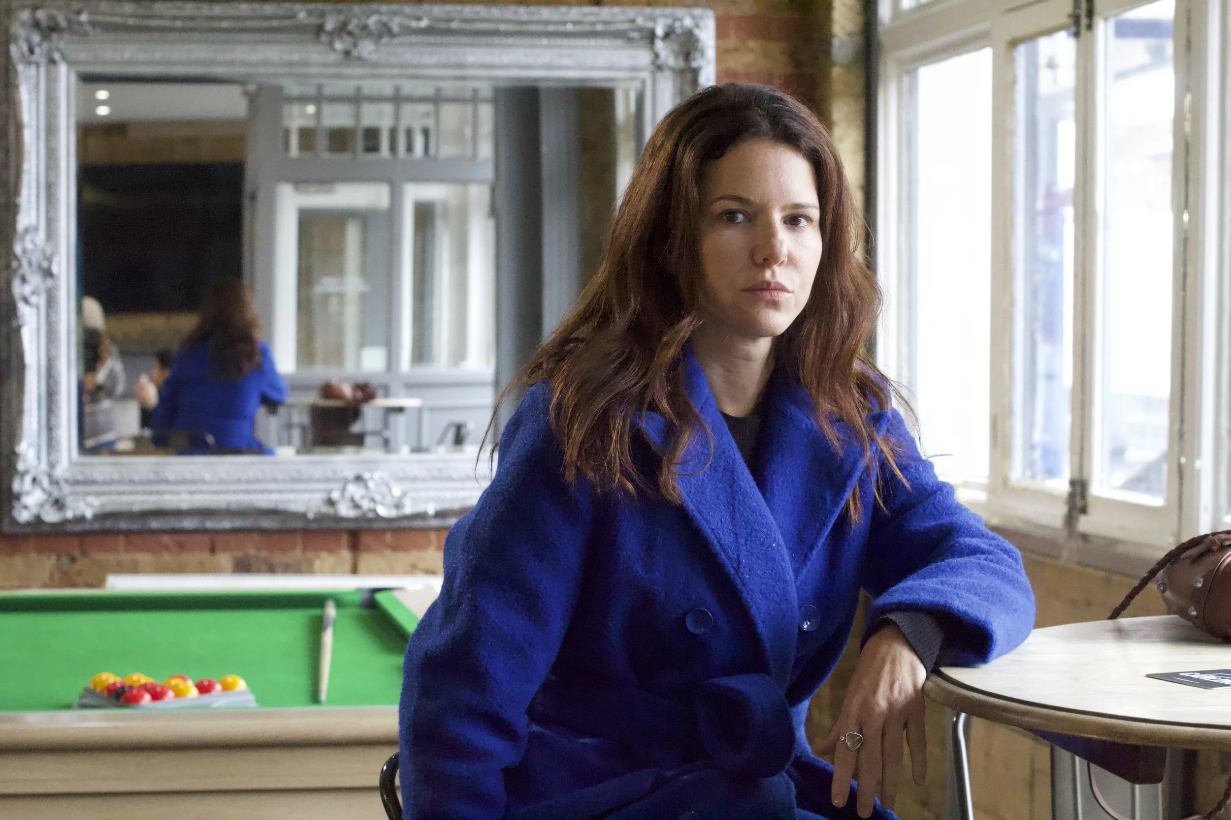 A woman with reddish-brown hair wearing a blue coat sitting at a table near a window in a bright room with large windows, a pool table, and brick walls.