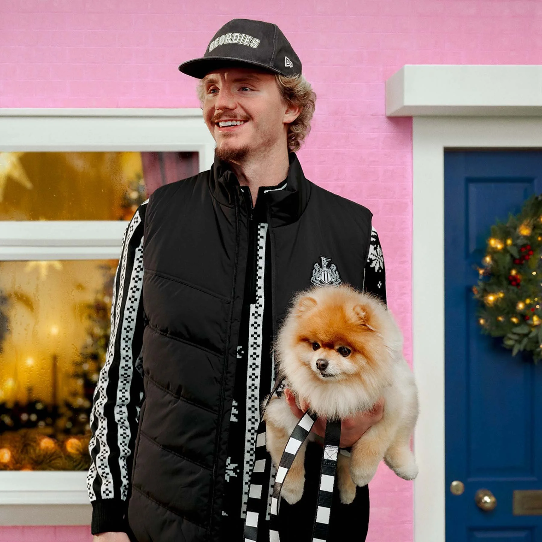 A man with blond hair and a beard, wearing a black cap and a black vest, holding a fluffy cream-colored dog in front of a colorful background.