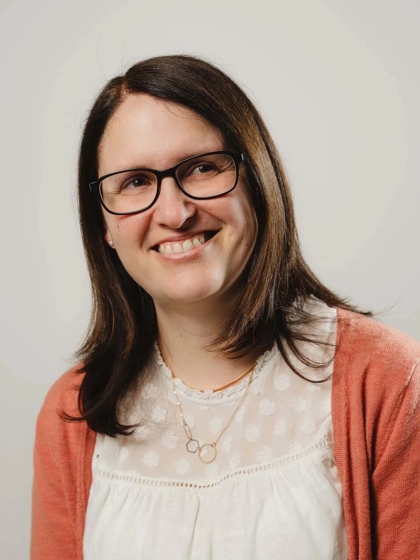 Close-up photo of a smiling woman with brown hair, wearing black glasses, a white blouse, layered necklaces, and an orange cardigan. White background.