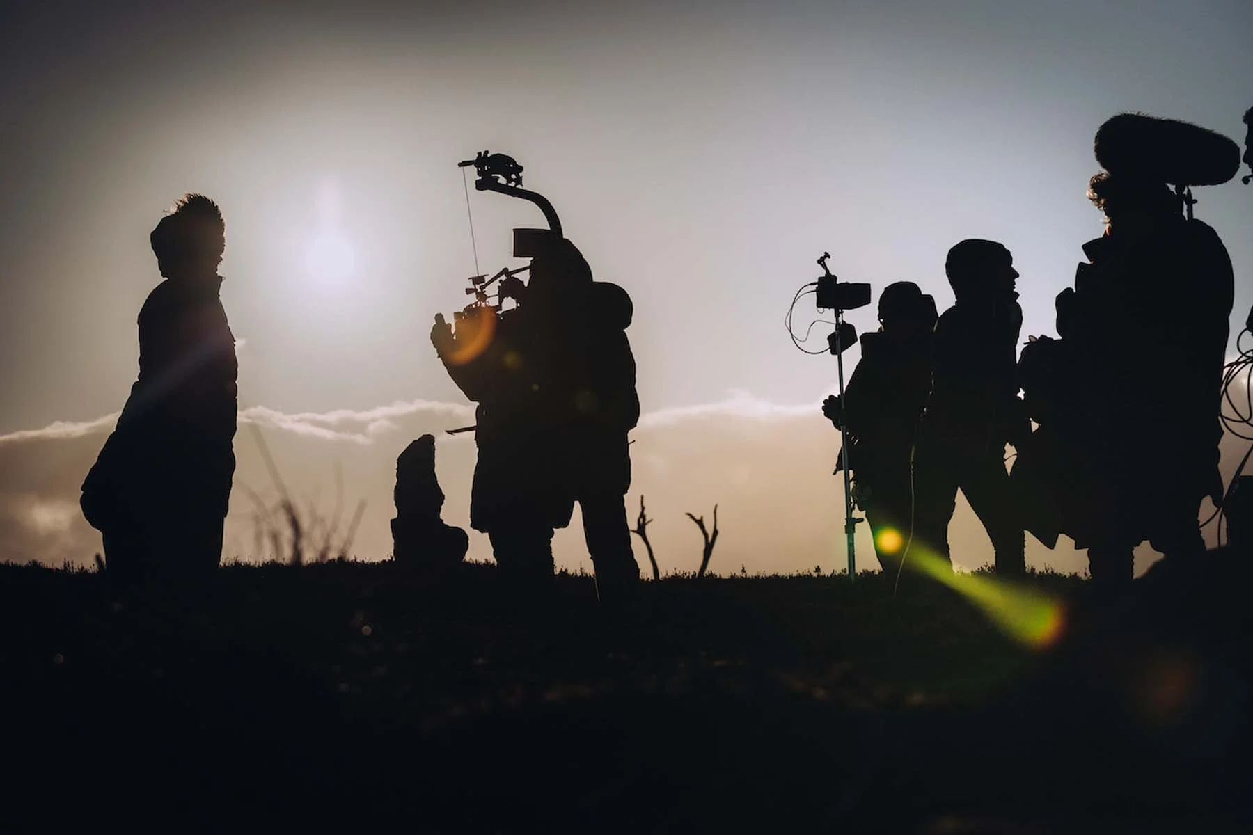 Silhouettes of people, including a child, standing outdoors during sunset, with some holding camera equipment, with a cloudy sky and a bright sun in the background.