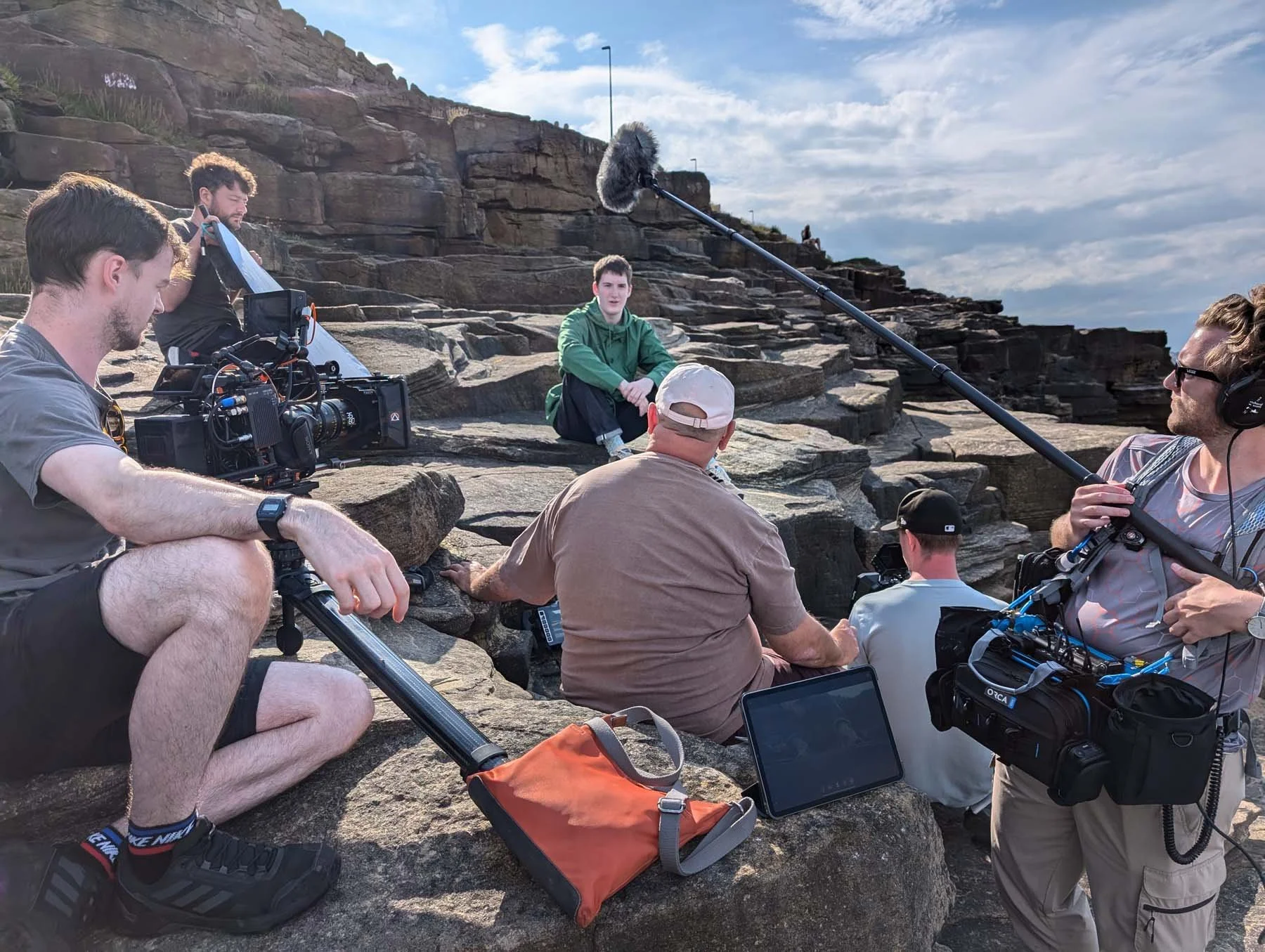 A group of people filming a young man sitting on rocks at a rocky outdoor location with a cloudy sky in the background. The young man is wearing a green hoodie and black pants, looking at the camera.