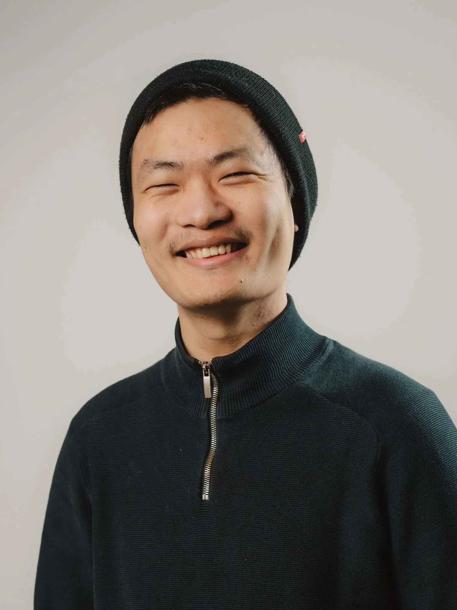A young man with short black hair, wearing a black knit beanie hat with a small red logo, and a black zip-up sweater, smiling against a plain light gray background.