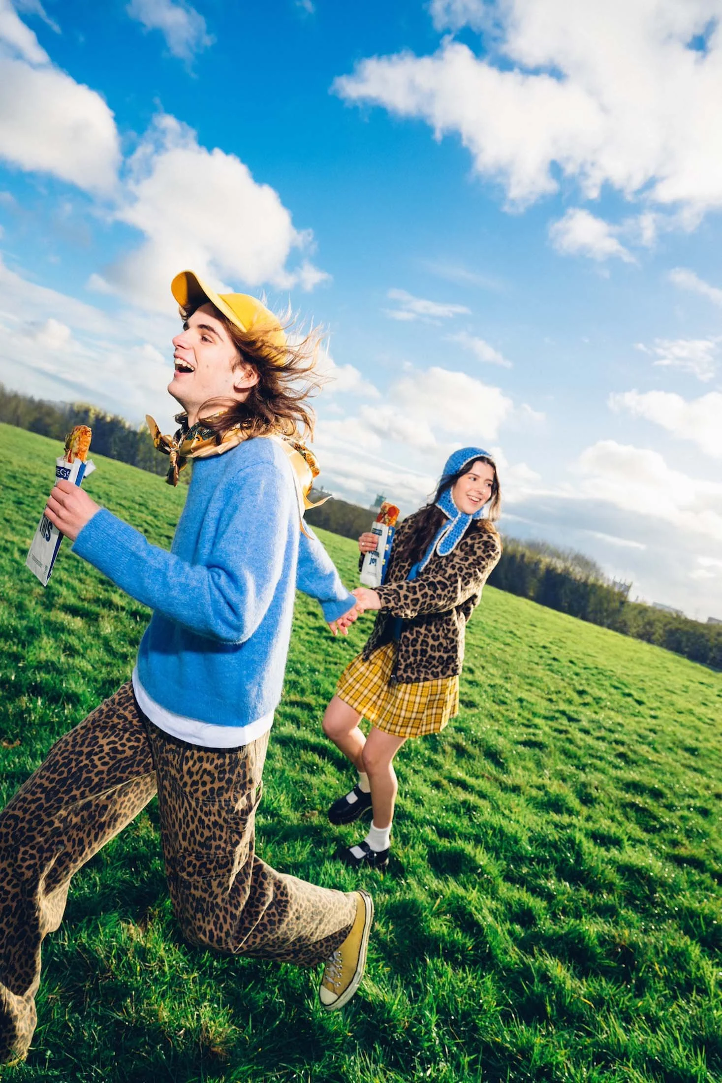 Two young people running and laughing through a grassy field on a sunny day. The person in front is wearing leopard print pants, a yellow cap, and a blue sweater, while the person behind is in a leopard print coat, yellow plaid skirt, and a blue knit hat, holding hands.
