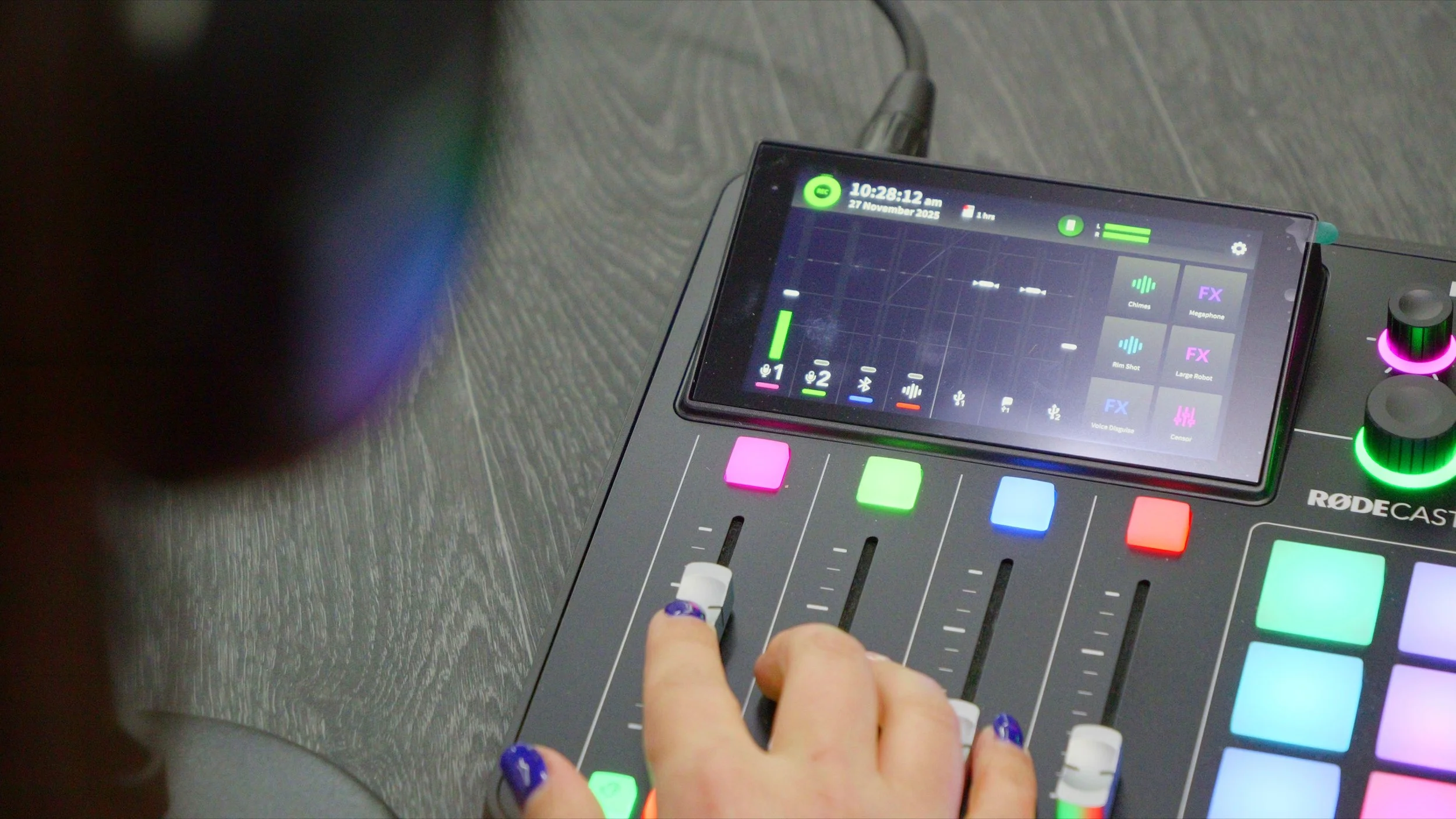 Close-up of a person using a RODECASTER audio control surface with a touchscreen interface displaying audio tracks and controls, placed on a black wooden surface.