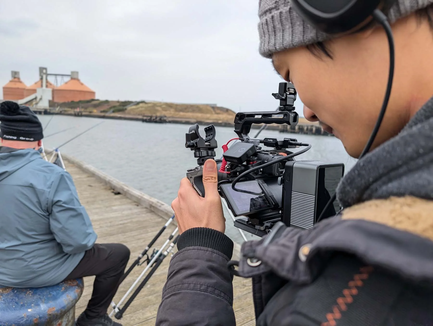Close-up of a person operating a professional video camera on a dock, with a man fishing in the background near water and buildings.