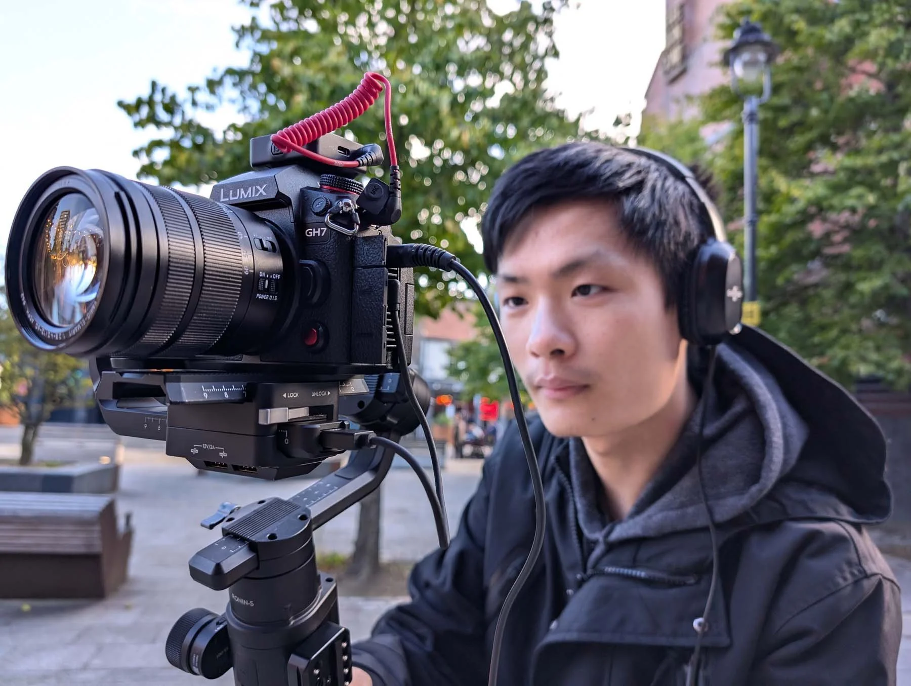 A young man wearing headphones operating a professional camera mounted on a stabilizer in an outdoor setting with trees and a building in the background.