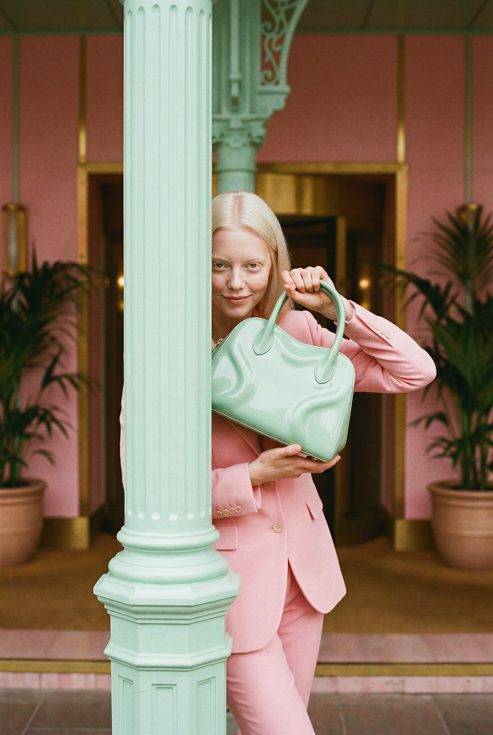 "Portrait of a model partially framed by a mint green architectural pillar, holding a matching handbag. An example of cinematic framing and high-end synthetic media production for luxury brands."