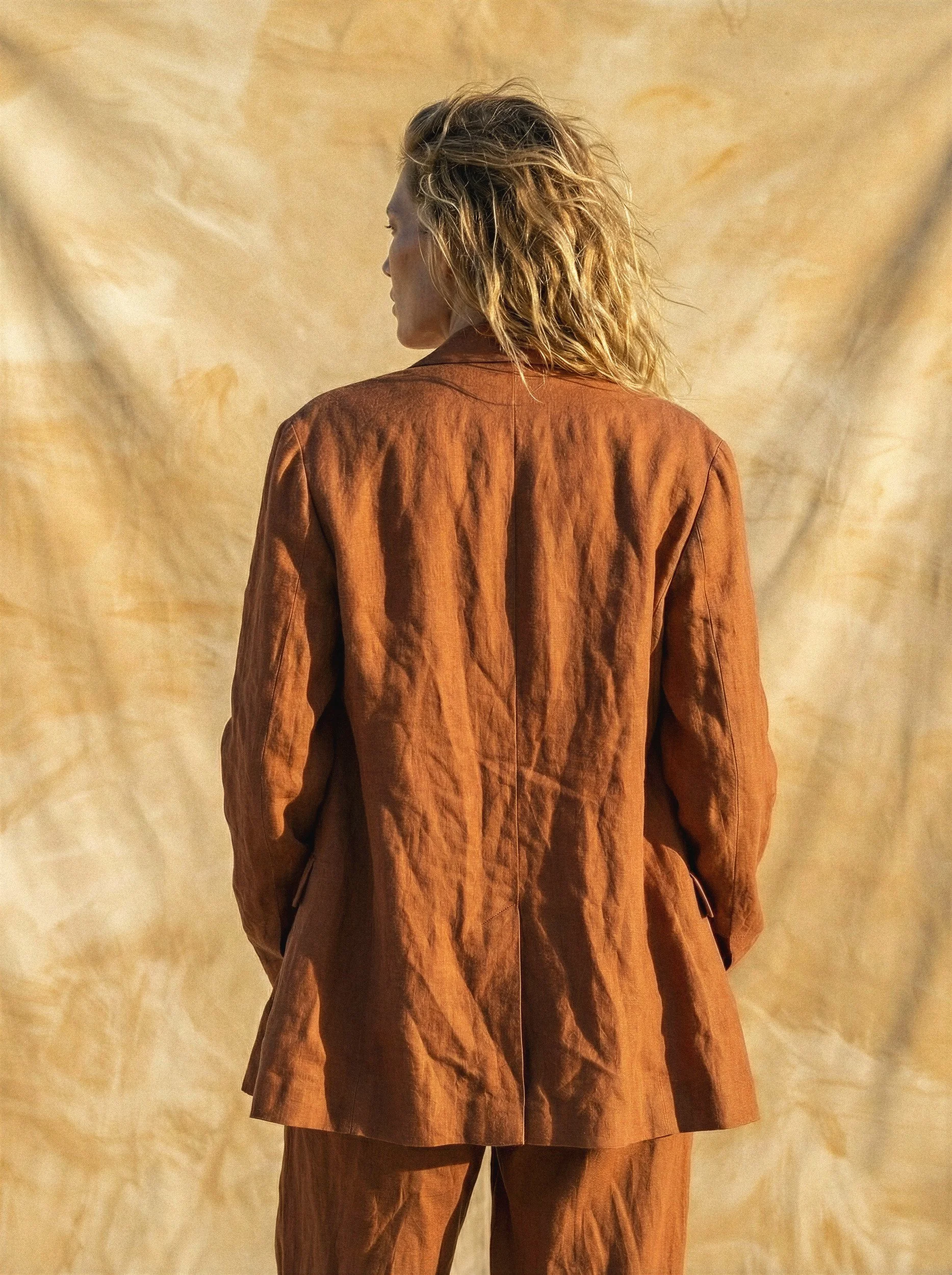 "Back view of a model in a tailored orange linen jacket standing against a sun-drenched textured fabric backdrop. Showcasing the precision of AI in rendering complex fabric folds and Mediterranean warmth for high-end fashion production."