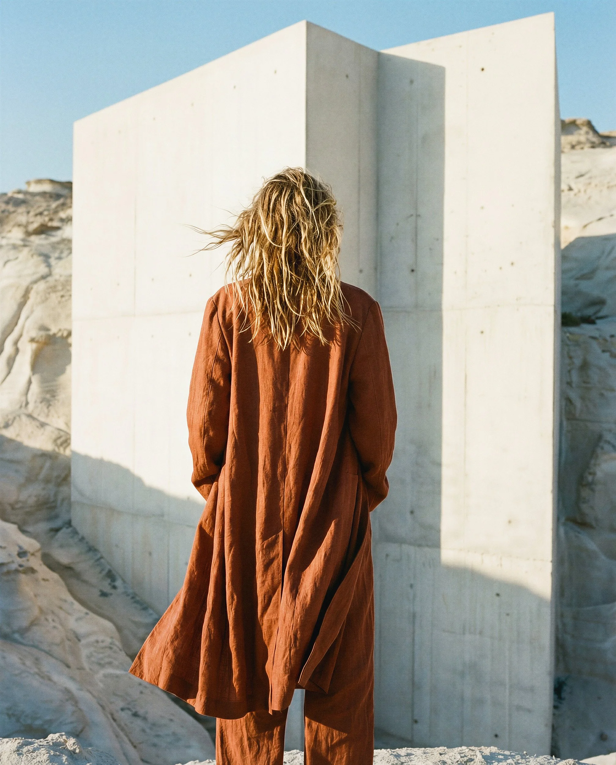"Back view of a model in an orange linen coat standing before a minimalist white monolith in a coastal setting. An exploration of volume, light, and synthetic architectural fashion photography."