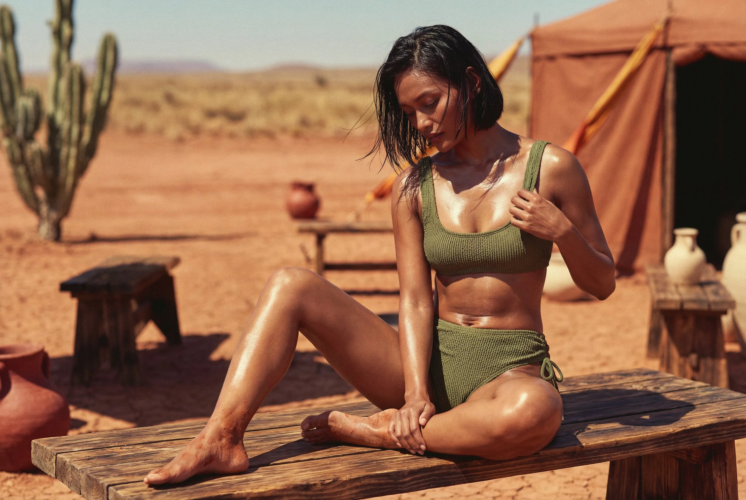 Cinematic desert photography of a model adjusting her olive green textured swimwear while sitting on a rustic wooden bench, surrounded by clay pots and dry earth under harsh sunlight.