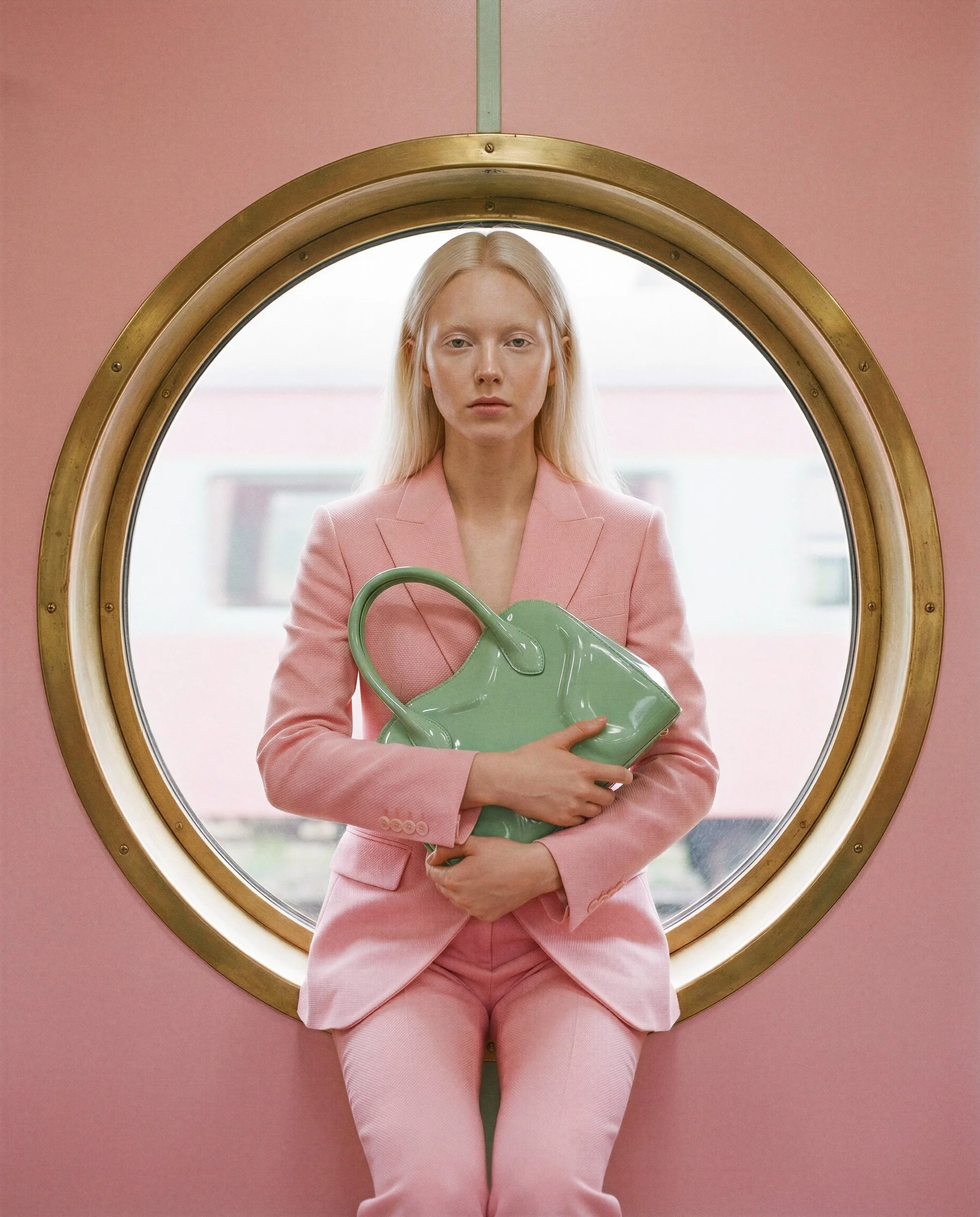 "Symmetrical portrait of a blonde model in a pink suit sitting centered within a circular window frame. Showcasing hyper-realistic fashion visuals and cinematic composition without physical sets."