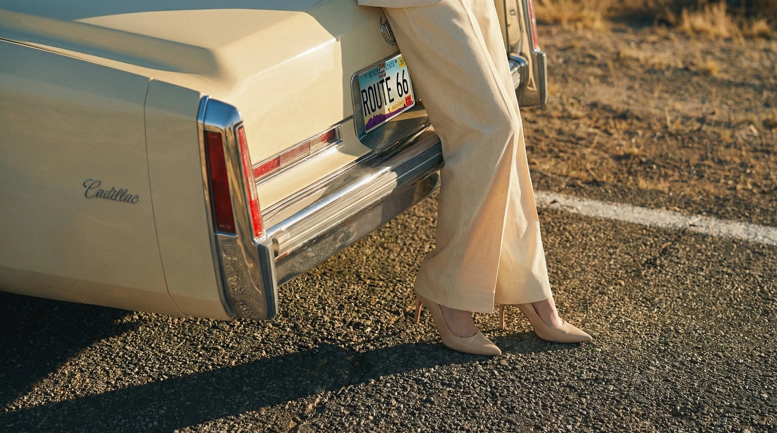 "Detail shot of a vintage car bumper with a 'Route 66' license plate, focusing on the model's cream trousers and beige heels. Demonstrating material realism and lighting accuracy in AI production."