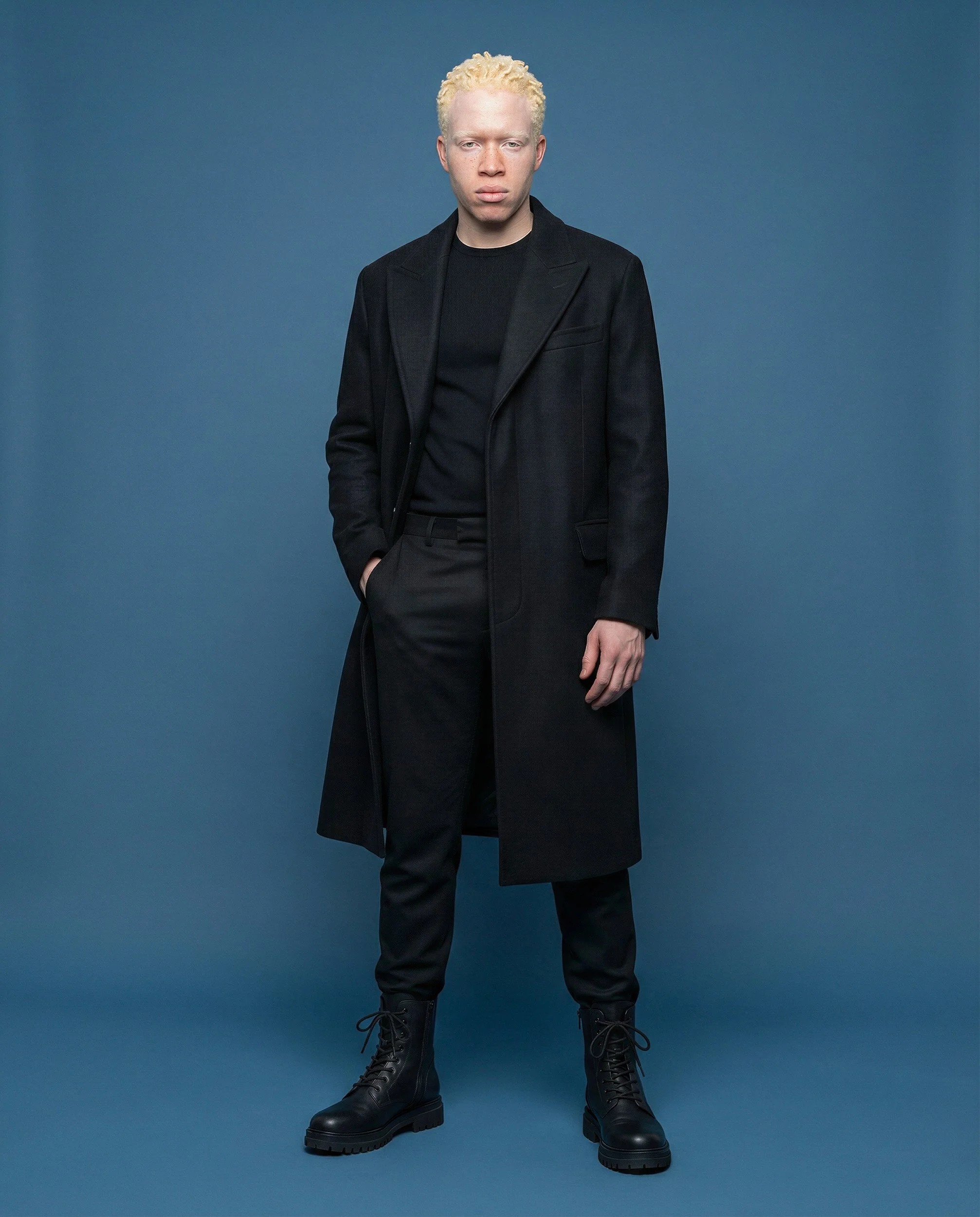 "Studio editorial portrait of a male model in monochromatic black tailoring against a deep blue background. Part of the Arctic Void campaign showcasing resilient tailoring and AI-generated fashion."