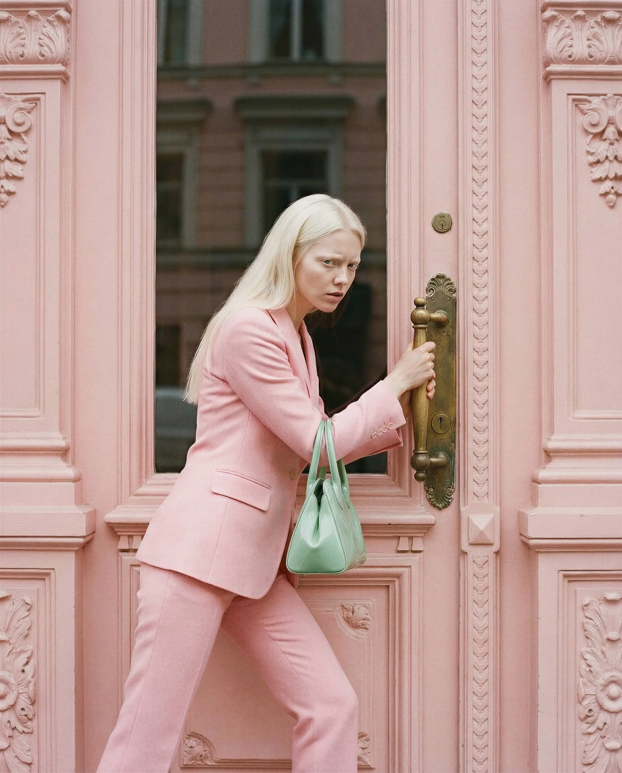 "Action shot of a fashion model in a pink suit holding a mint bag while opening an ornate pink door. Illustrating AI-generated fashion realism and dynamic human interaction with synthetic architectural elements."