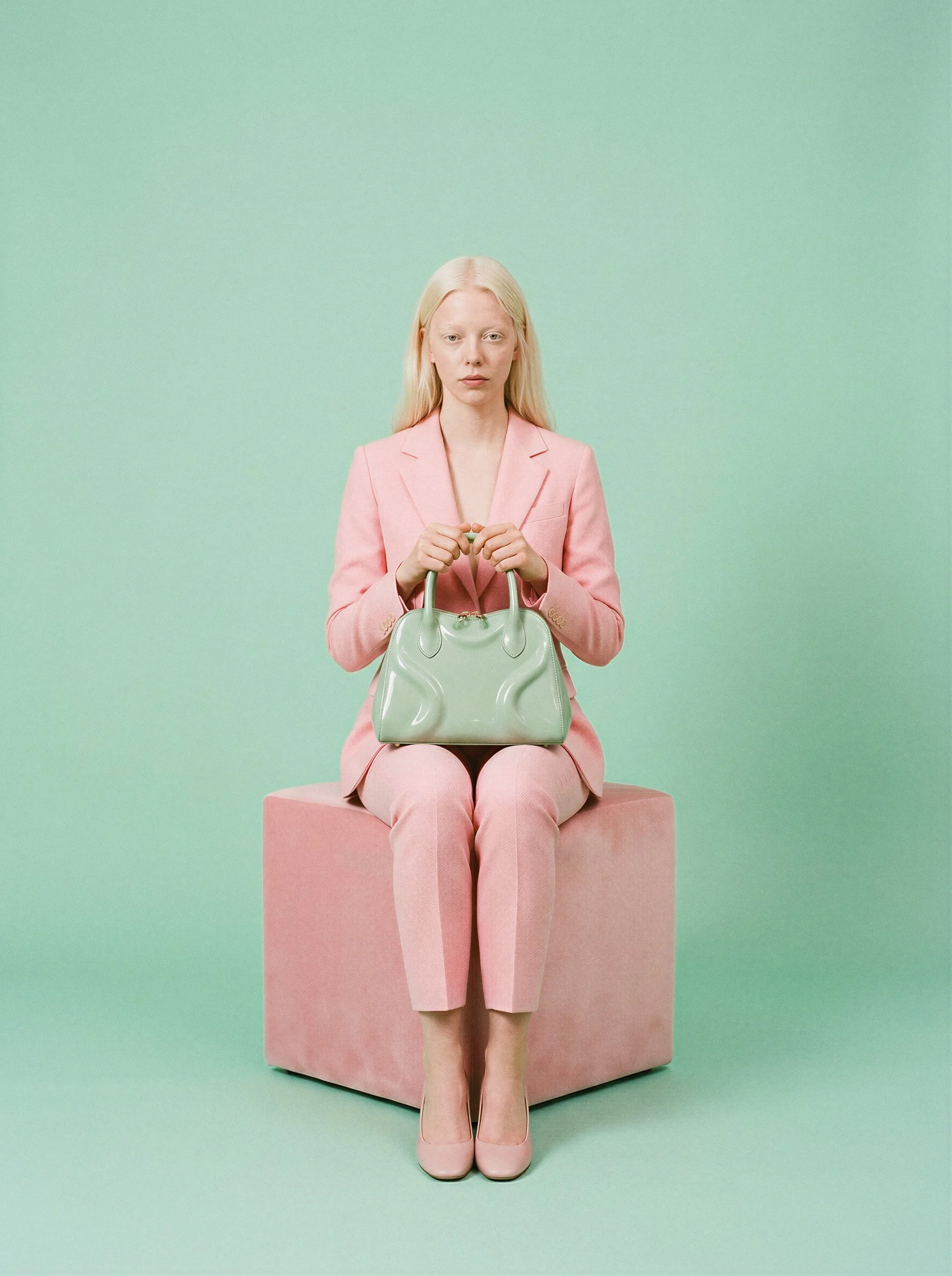"Symmetrical studio portrait of a model sitting on a pink velvet cube holding a mint green bag against a solid mint green background. Showcasing the clean, high-fidelity aesthetic of zero-logistics AI fashion production."
