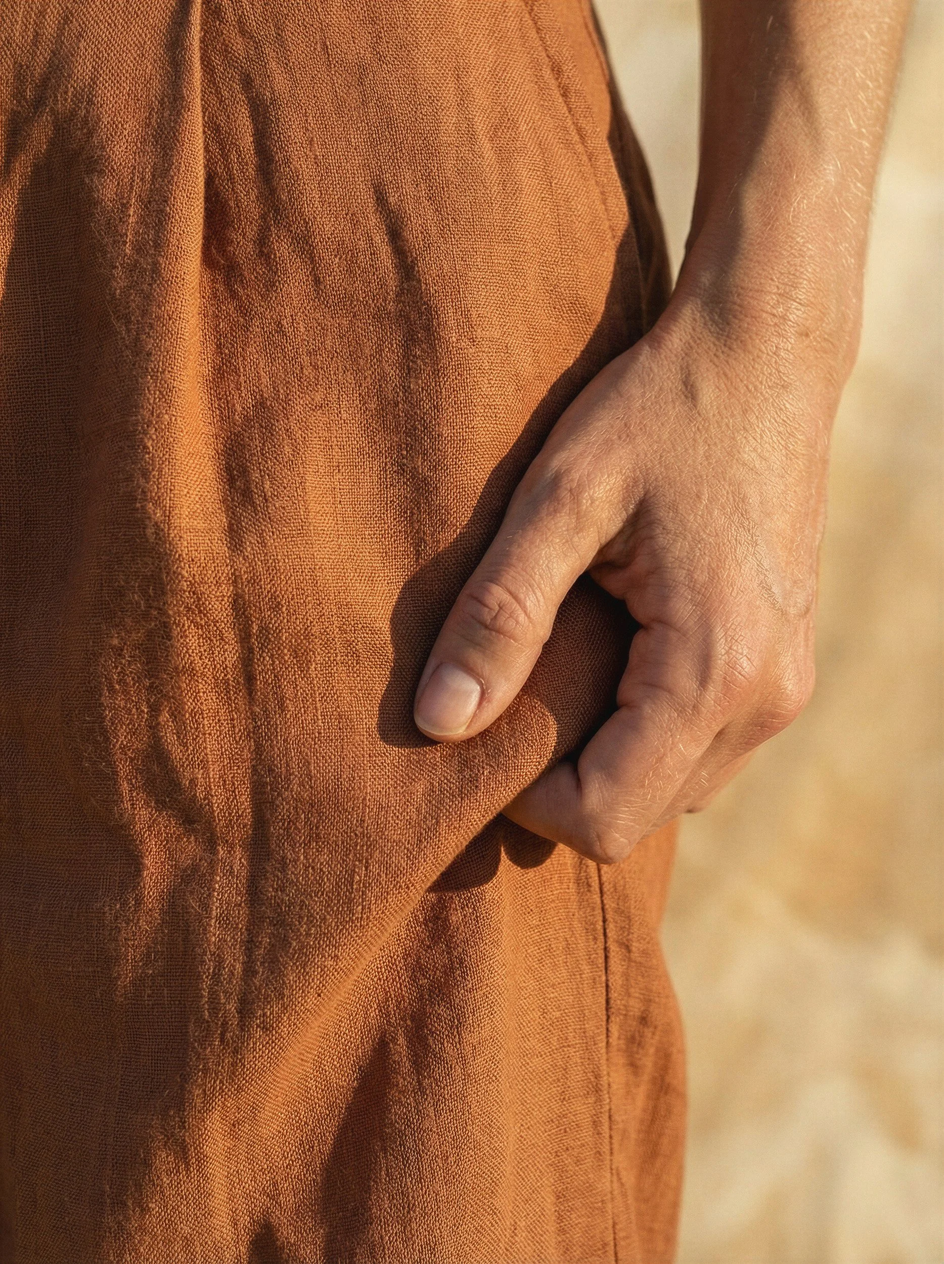 "Extreme close-up detail of a hand resting in the pocket of terracotta linen trousers. Highlighting the hyper-realistic weave, organic texture, and sunlit skin fidelity achieved by The Raw Lab's advanced AI visual engine."