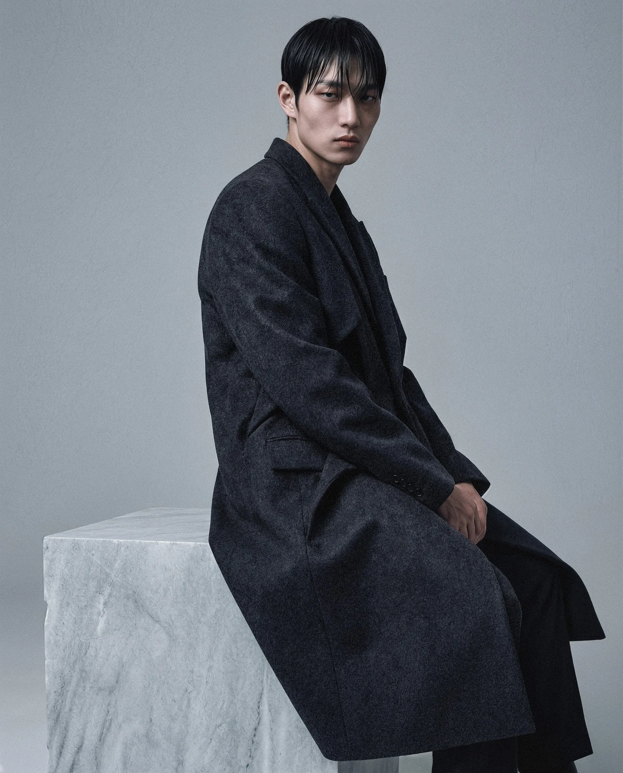 "A digital twin male model in a structured black wool coat sitting on a large white marble block. Studio shot for the Monolith campaign by The Raw Lab, demonstrating precision tailoring and AI visual engineering in Lisbon."
