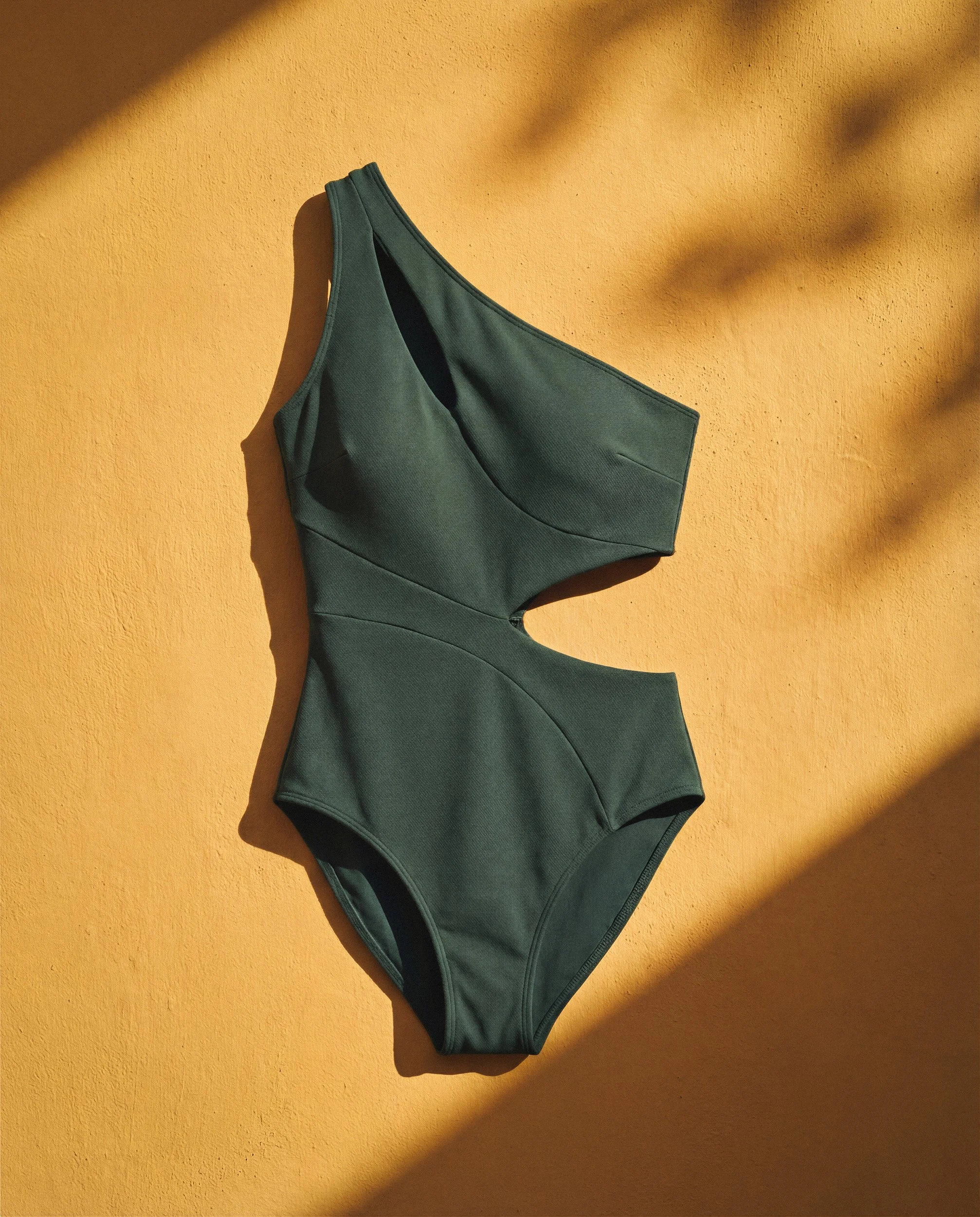 High-fashion product flat lay of a forest green cutout one-piece swimsuit on a weathered ochre background with natural sunlight and palm shadows.