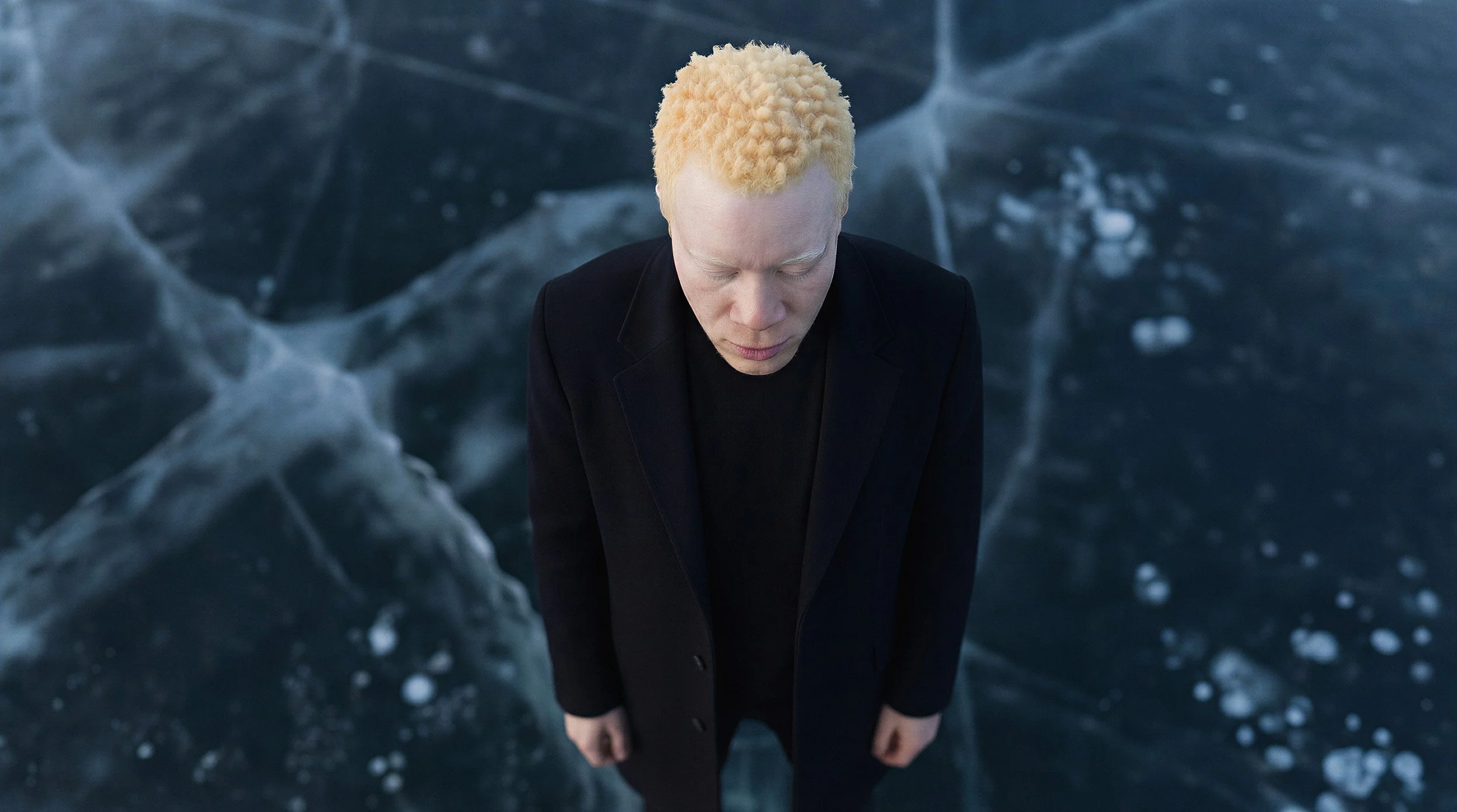 "High-angle overhead view of a model in a black coat standing on a geometric pattern of cracked ice. A study in sub-zero contrast and monochromatic fashion silhouettes."