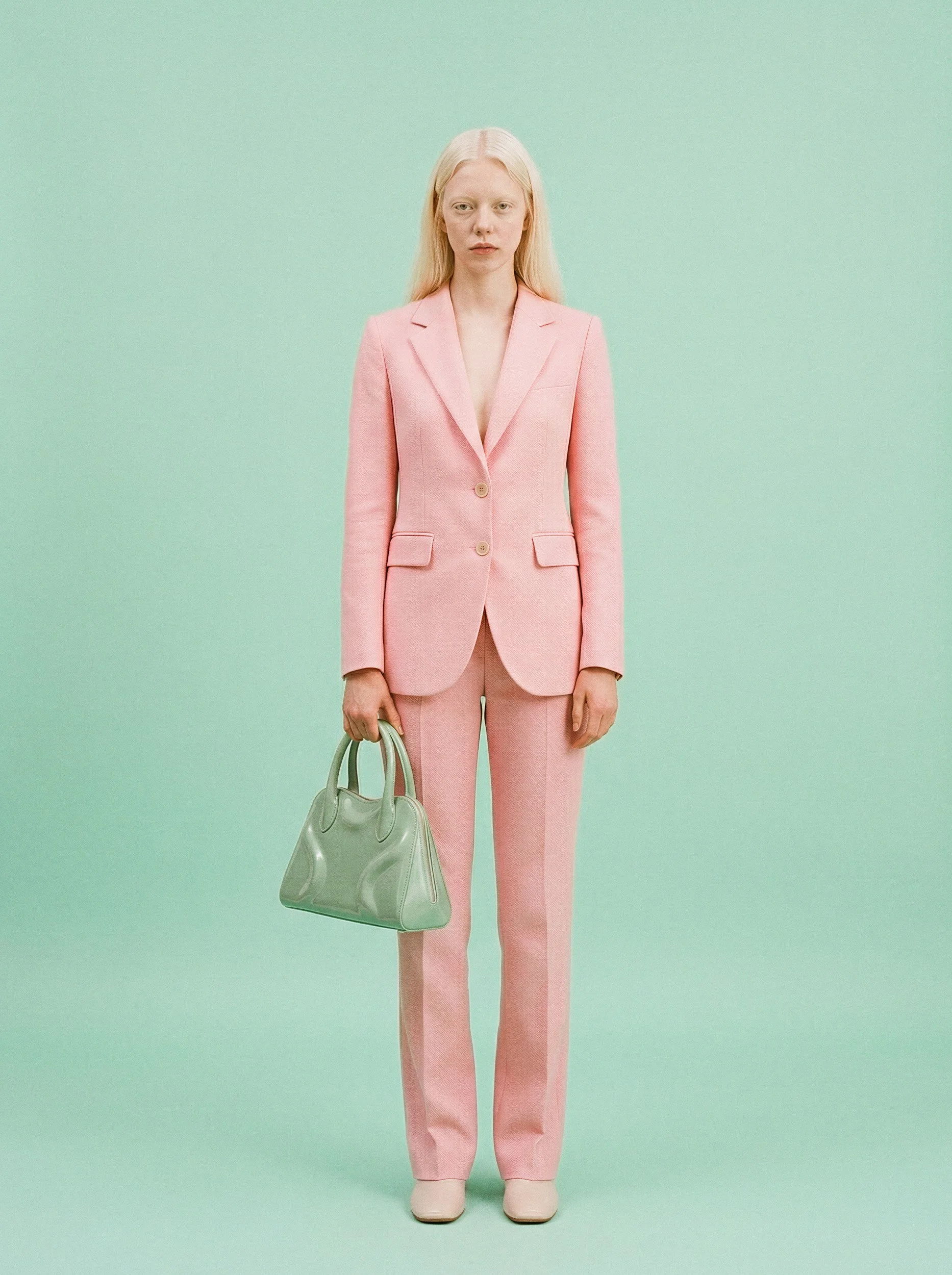 "Full body studio portrait of a digital twin model wearing a tailored pink suit and holding a mint green handbag against a solid mint green background. High-fidelity synthetic fashion visuals from the Symmetrical Utopia campaign by The Raw Lab."