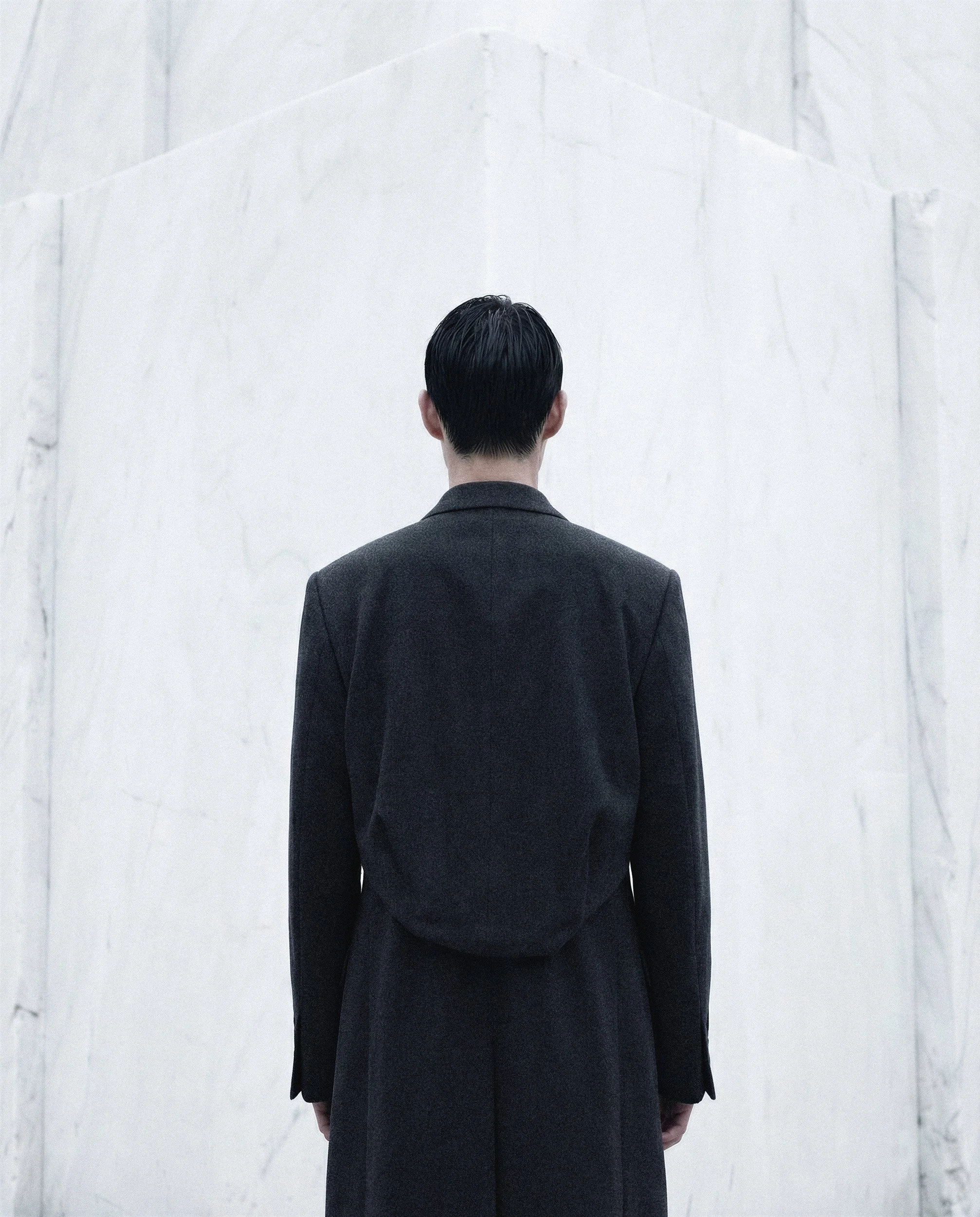 "Back view of a structured black wool coat against the geometric architecture of a marble quarry. A study in volume and monochromatic fashion silhouettes for the Monolith project."