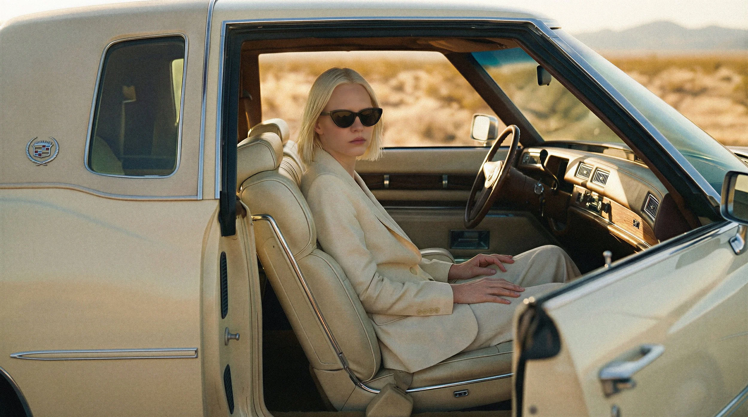 "Model in a cream suit sitting inside a vintage Cadillac with beige leather interior. Cinematic lighting through the window captures the desert mirage atmosphere without physical on-location logistics."