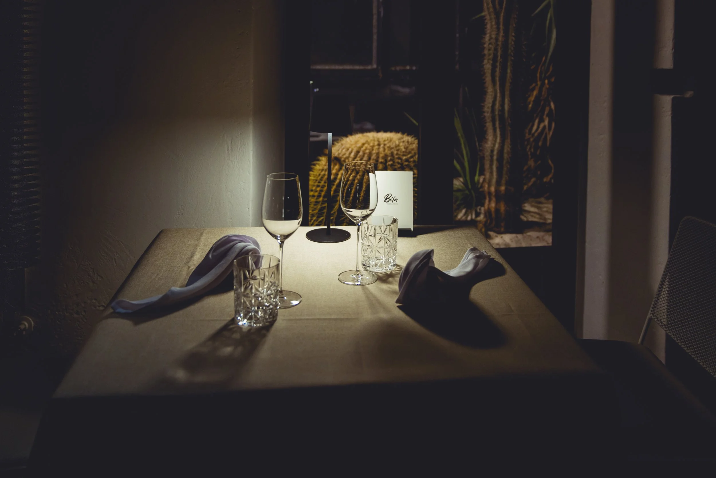 A dimly lit dining table set with wine glasses, water glasses, napkins, and a small menu card in a restaurant or cafe, with a cactus visible through the window in the background.