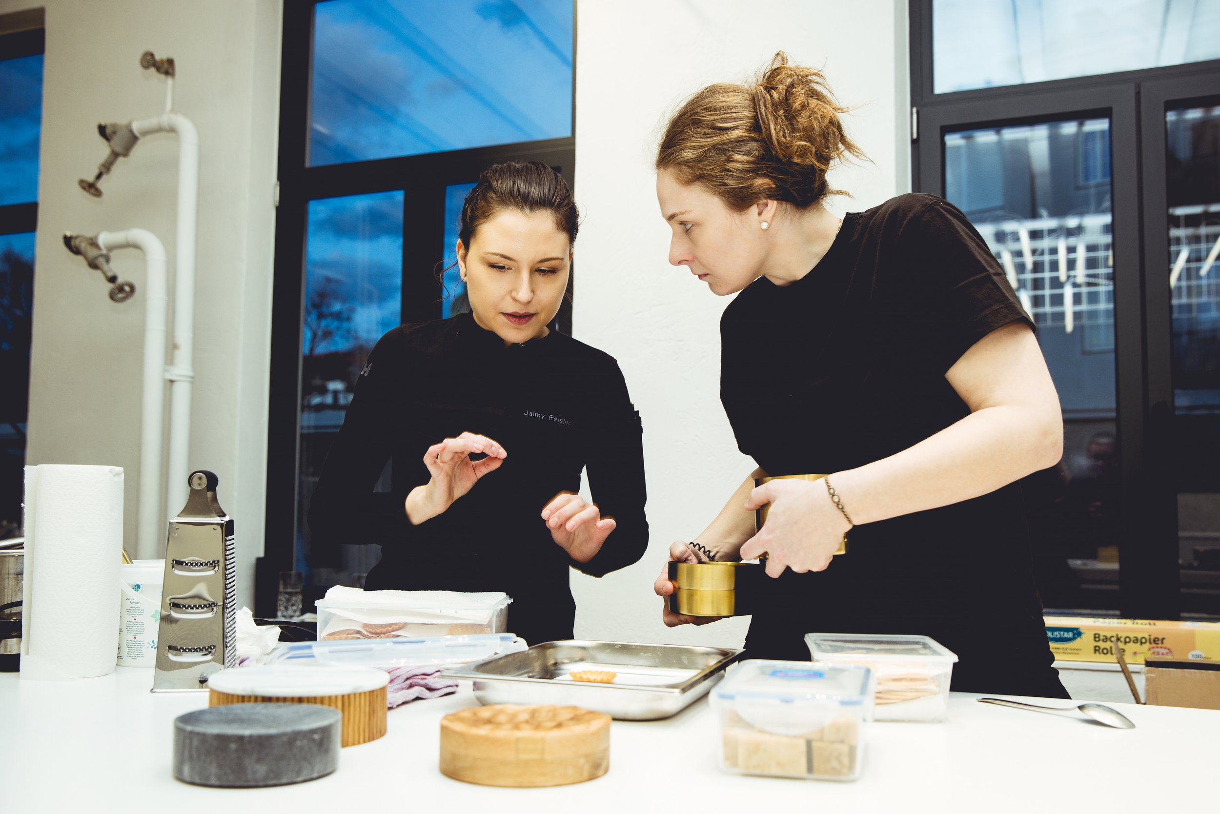 Jaimy Reisinger and Kathi Diwald are working together in a kitchen or bakery, preparing food with various kitchen tools and containers on the table.