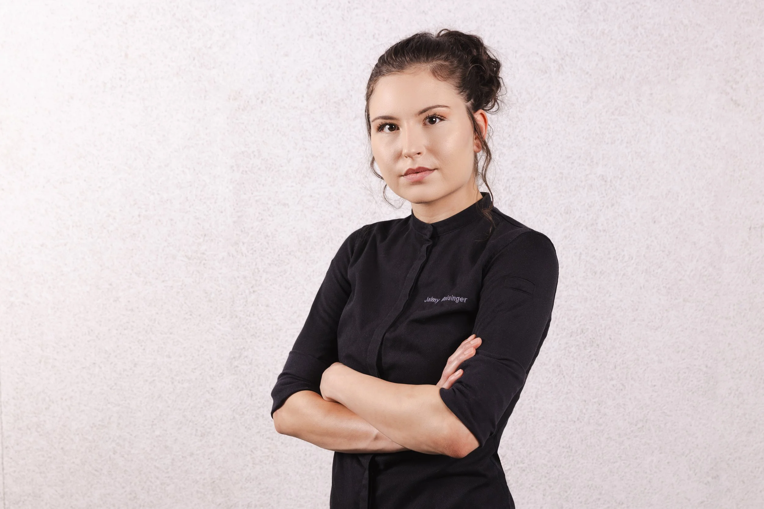Jaimy Reisinger with dark hair pulled back, wearing a black chef's coat with her arms crossed, standing against a plain, light-colored background.