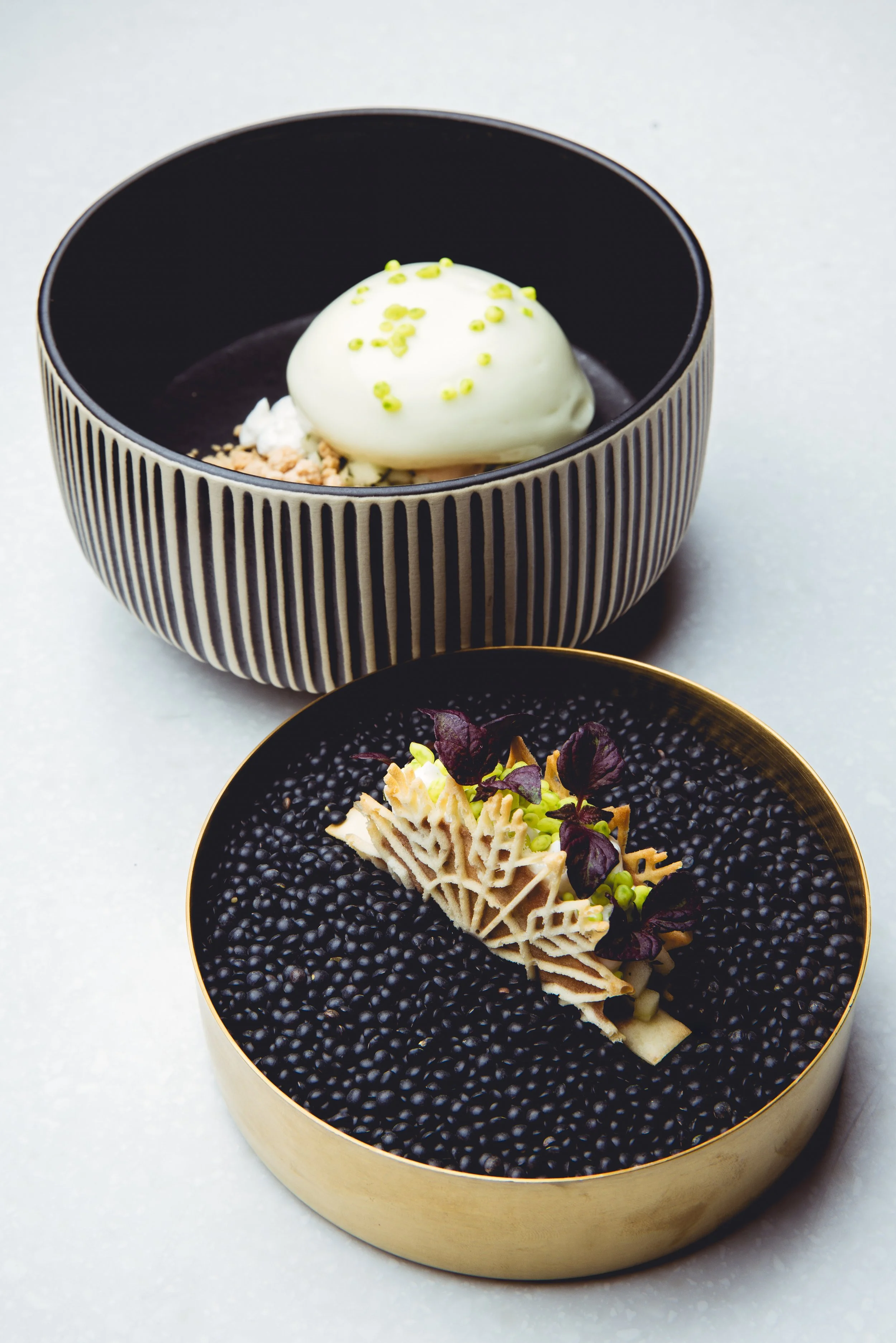 Two elegant plated desserts on a white surface: one with white chocolate mousse garnished with pistachio bits, the other with black sesame seeds and decorative chocolate lattice by Jaimy Reisinger