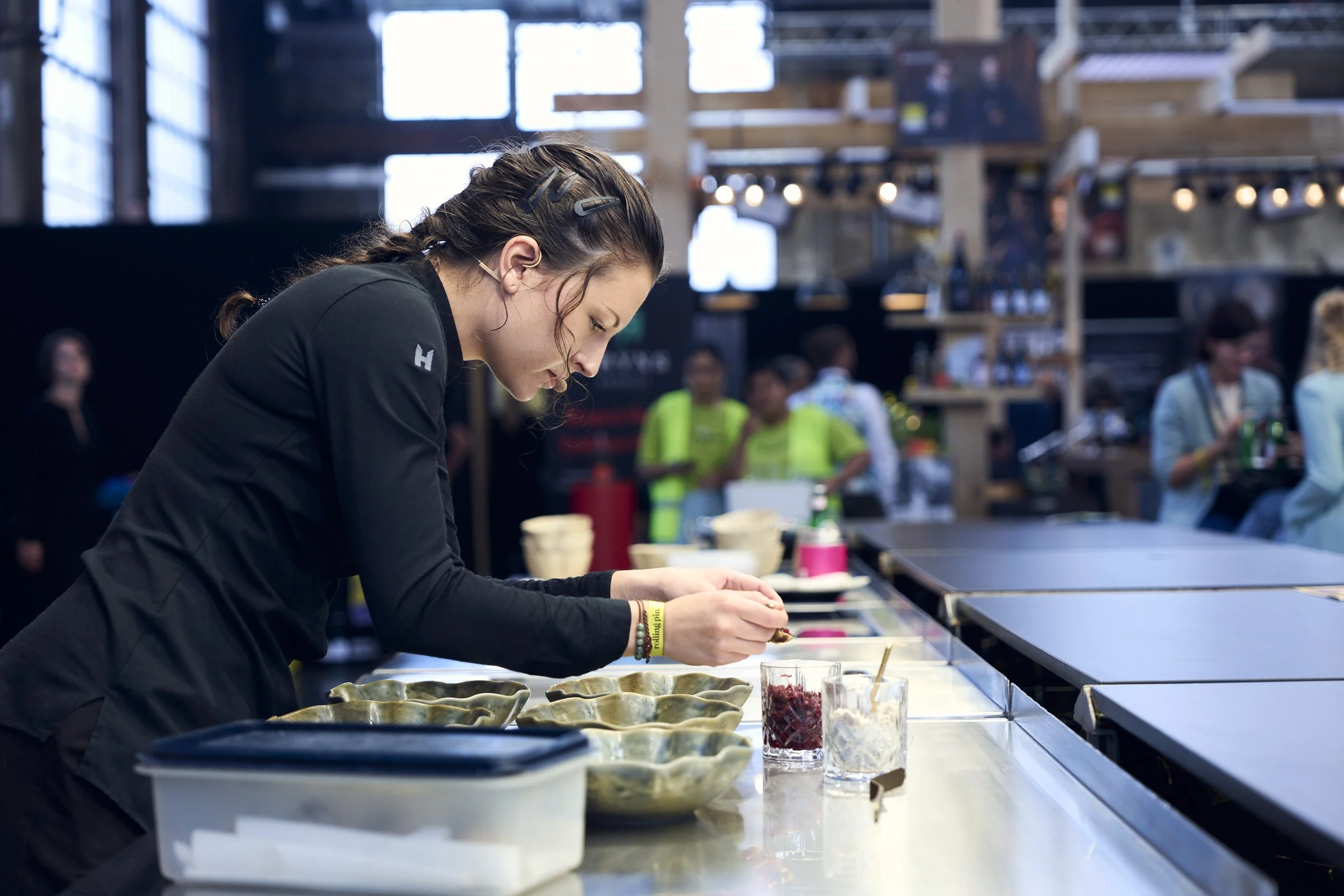 Jaimy Reisinger wearing a black jacket, preparing food at a counter in a busy indoor market or food event, with several other people in the background.