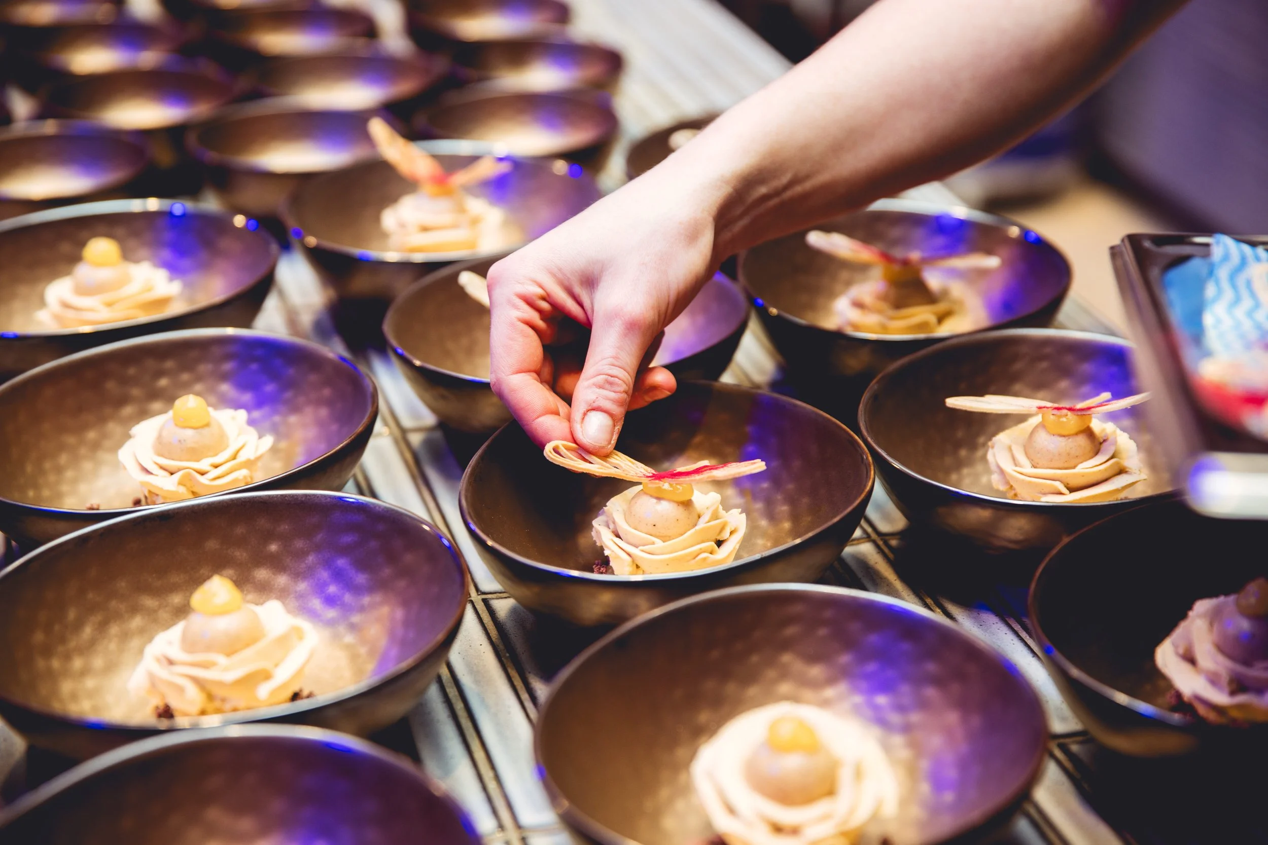 Jaimy Reisinger places an appetizer on a dark bowl, with many similar bowls arranged on a metal surface, all containing a small pastry topped with a dollop of yellow sauce and a thin crispy garnish.