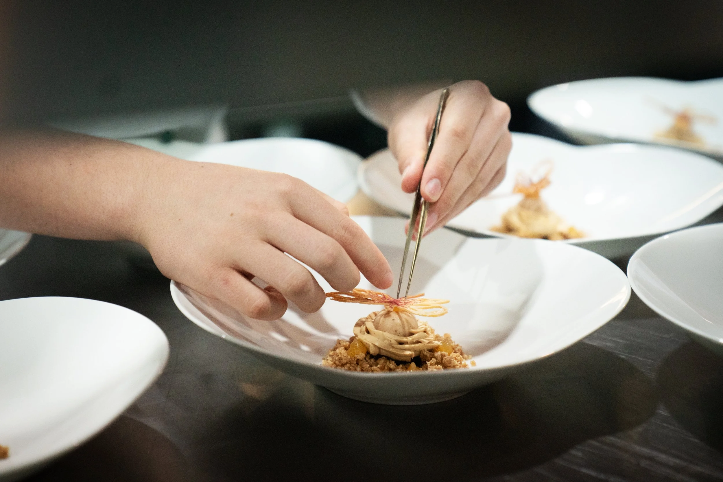 Chef carefully arranging a dessert with tweezers on a white plate, surrounded by more plates with similar desserts on a dark table.